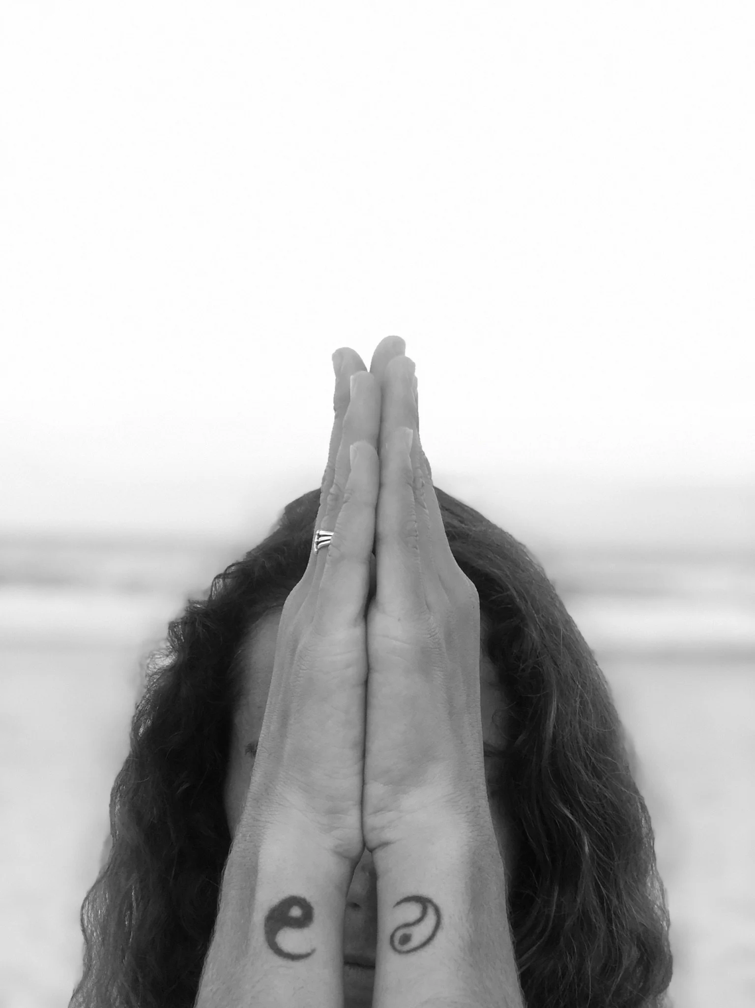 A person with long curly hair holds their hands together in a prayer position covering their face, with visible tattoos on their wrists, against a blurred outdoor background.