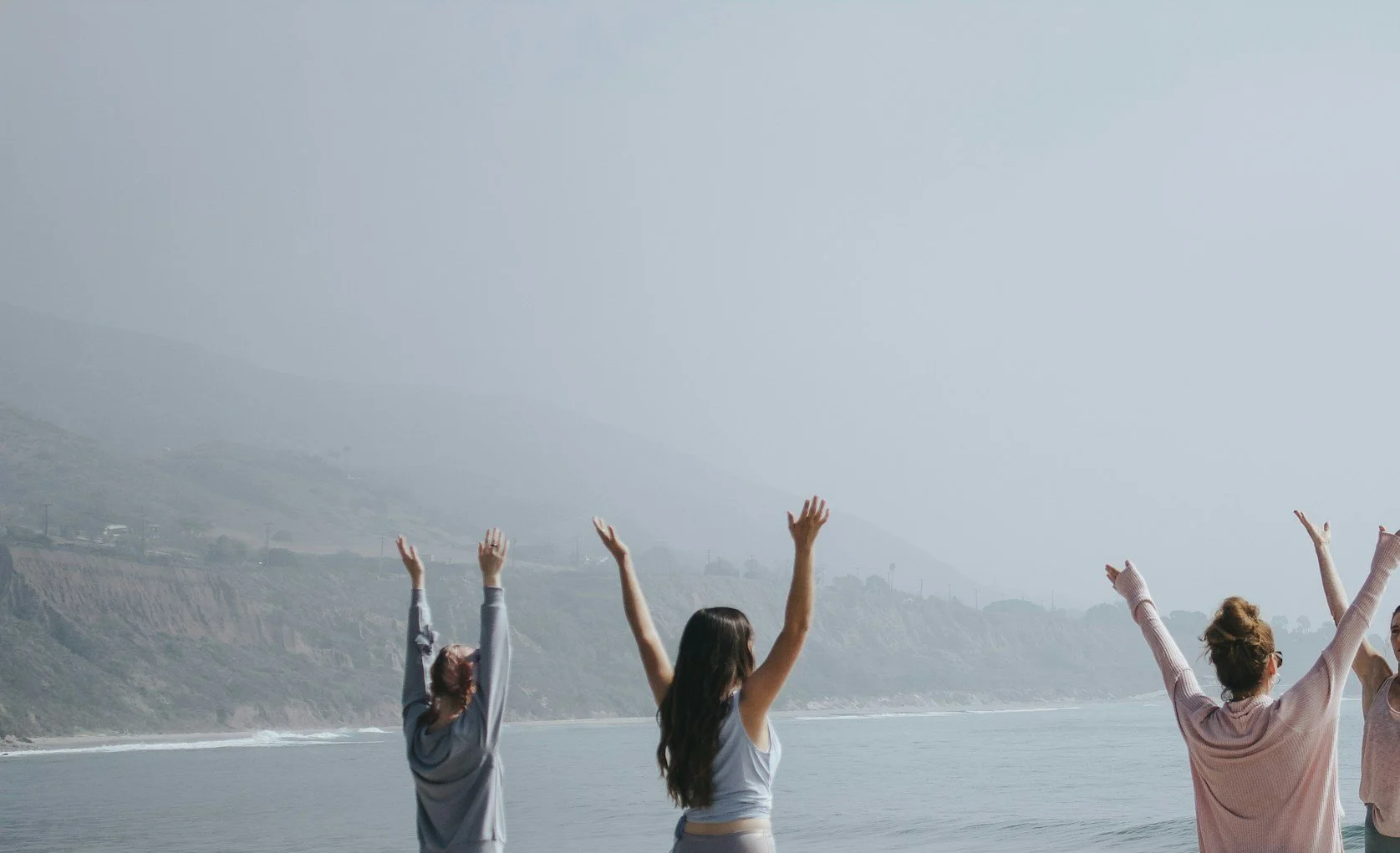 Four women standing on the shoreline with arms raised, facing the ocean, with a foggy or hazy sky and cliffs in the background.