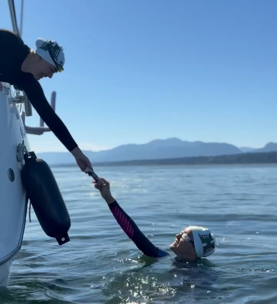 Person in the water reaching up to a person on a boat, with mountains in the background, during daytime.