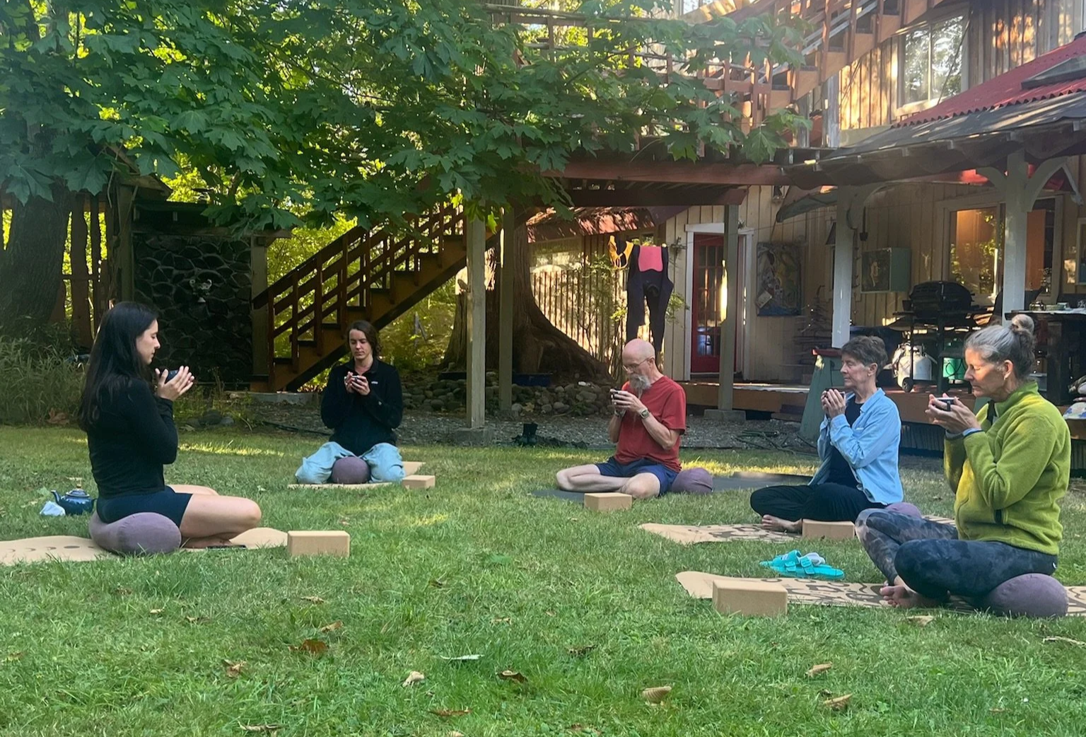 Group of five people practicing meditation outdoors on grass, seated on cushions, with each person holding a smartphone, in a backyard with trees, wooden house, and outdoor stairs.