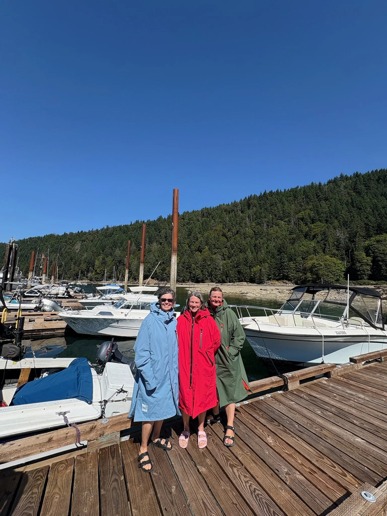 Three women standing on a wooden dock with boats and a forested hillside in the background on a sunny day.