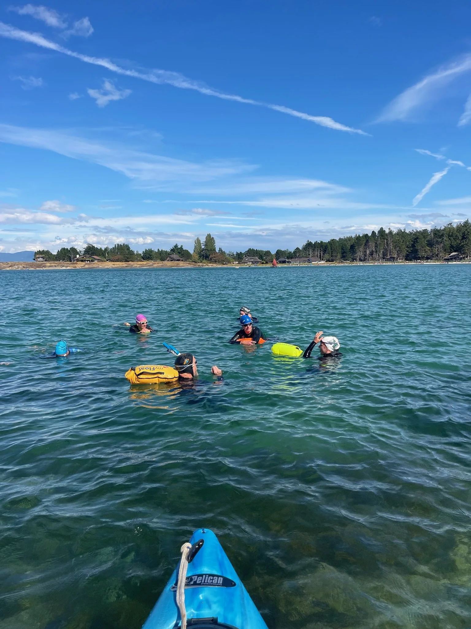 People in life jackets and wetsuits swimming and floating in a lake, with a blue sky and scattered clouds above.