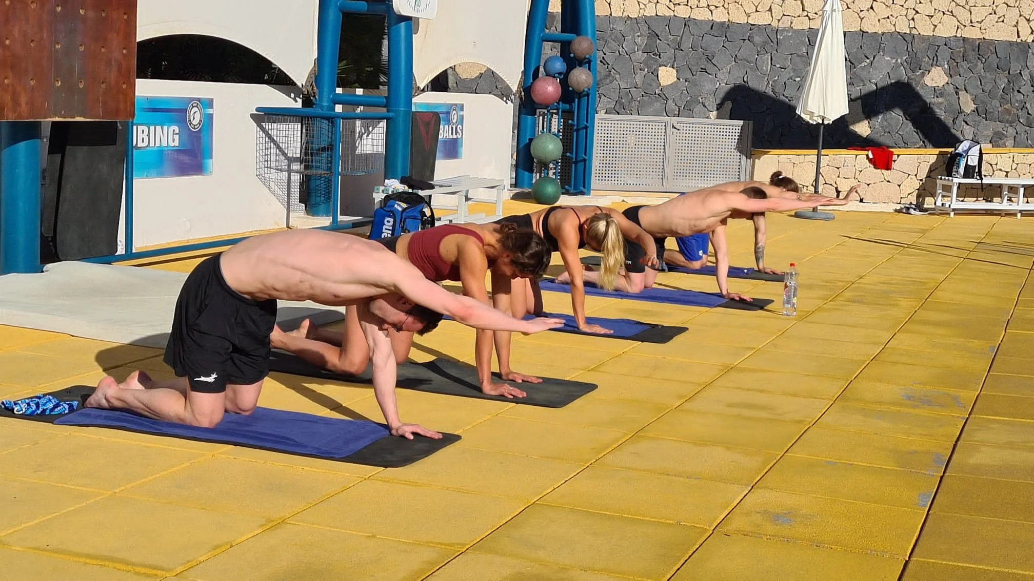 Four people practicing yoga outdoors on yellow mats, in a downward extended position, with one arm and opposite leg extended, on a yellow surface with a stone wall and patio umbrella in the background.
