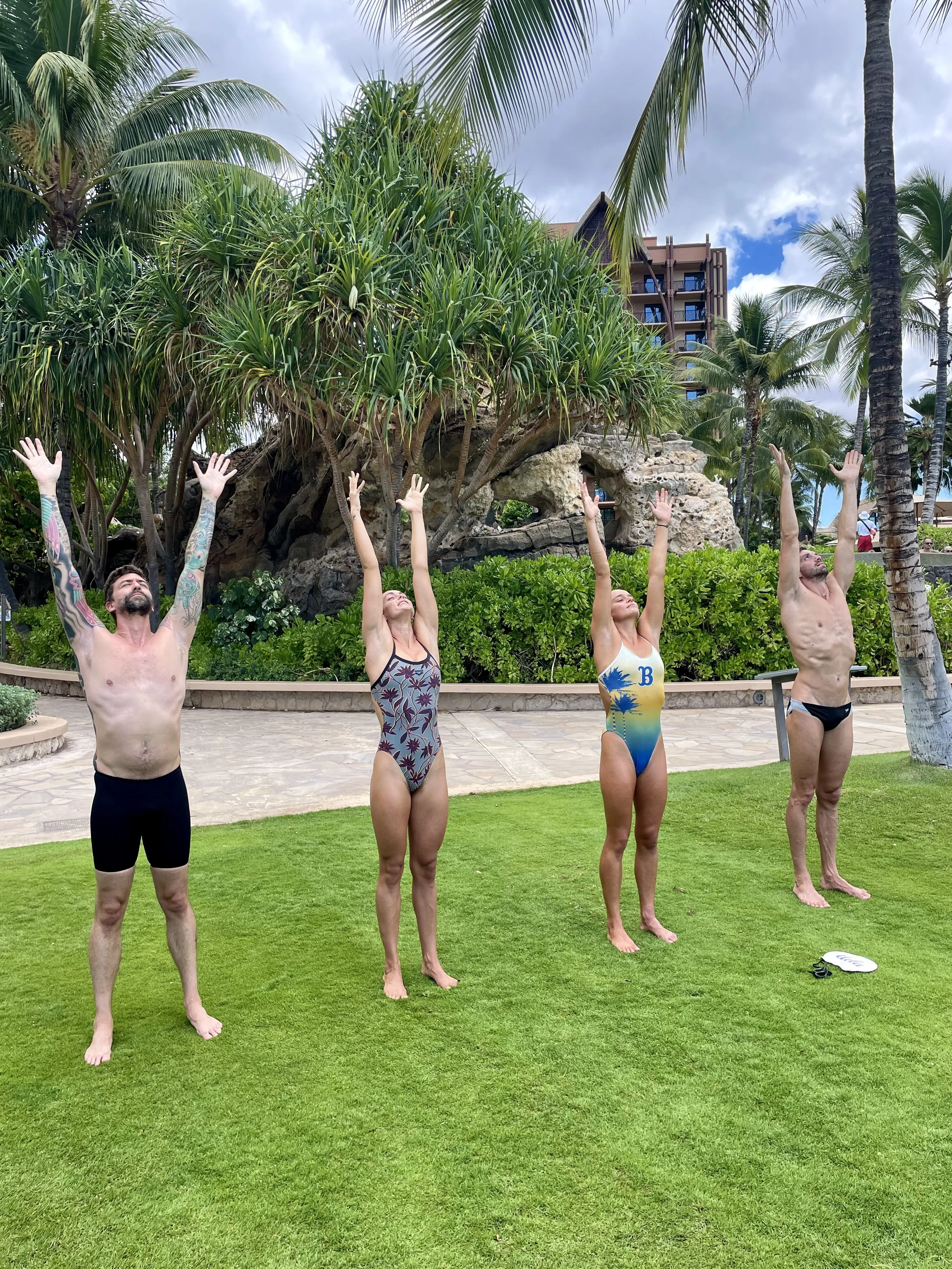 Four people standing on grass with arms raised in a yoga pose, outdoors at a tropical location with palm trees and lush greenery.