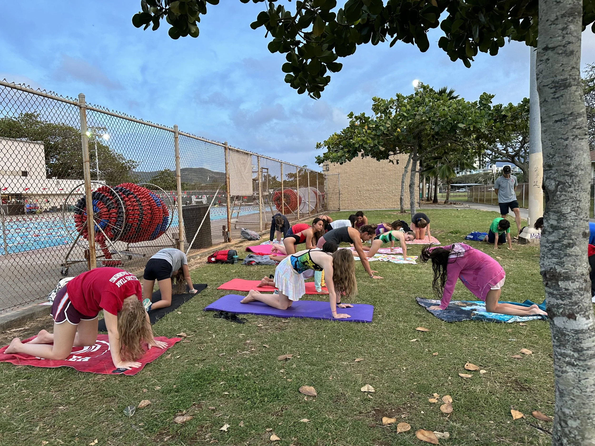 Group of people practicing yoga outdoors on mats near a swimming pool, under trees, with a chain-link fence, pool equipment, and a person standing nearby.