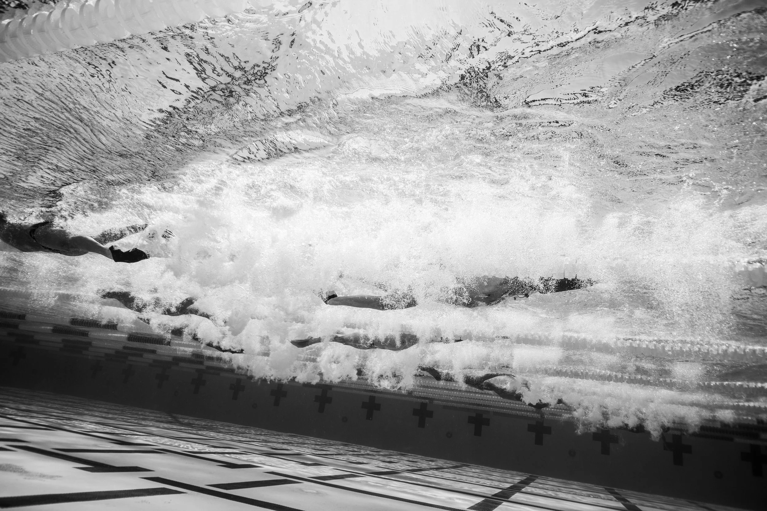 Swimmers in an indoor pool, captured from beneath the water surface, with bubbles and water movement.