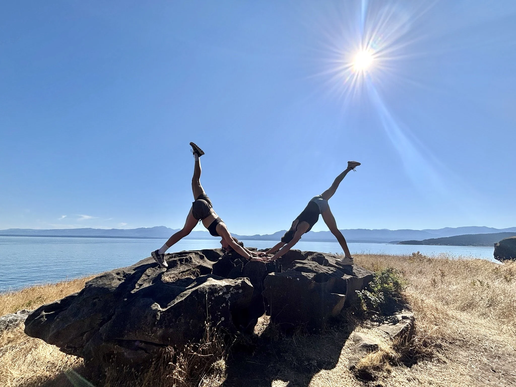 Two women in athletic clothing perform a yoga pose on a large rock by a body of water under a bright sun in a clear sky.