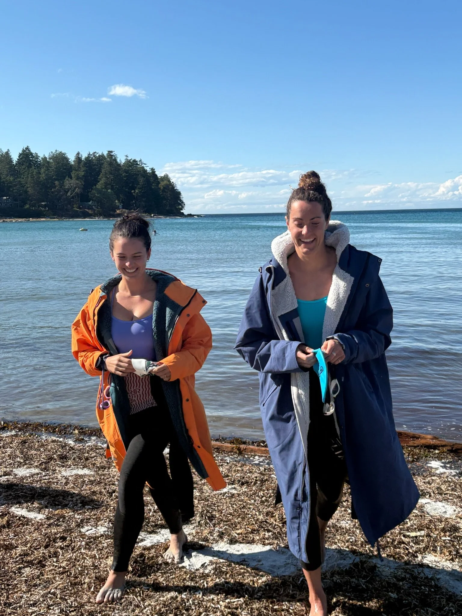 Two women in jackets walking barefoot on a beach near a body of water, smiling and holding items, with trees and a partly cloudy sky in the background.