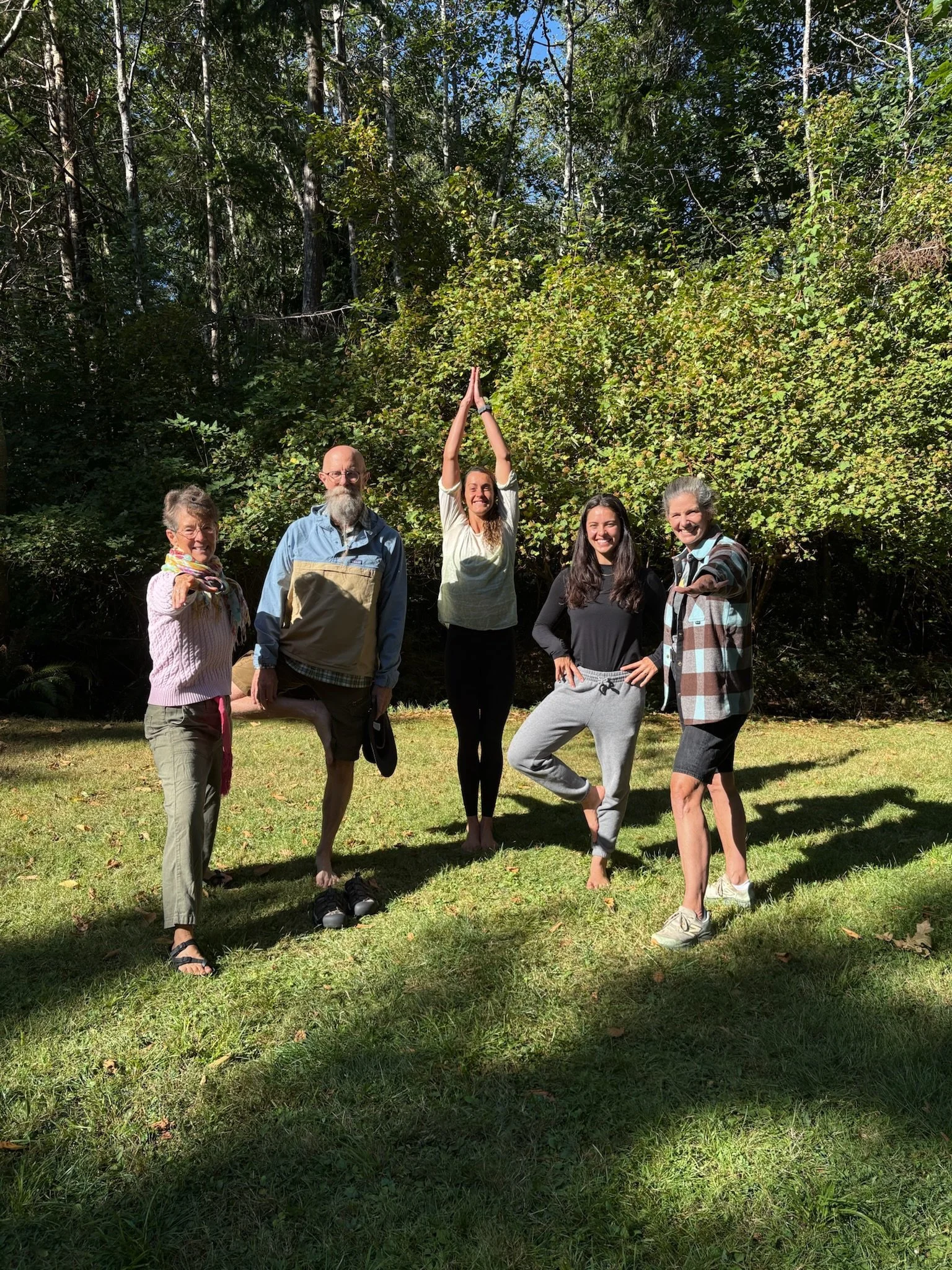 Five people standing outdoors on grass, with trees and bushes behind them. They are posing, some smiling directly at the camera. One woman in the middle has her arms raised above her head in a prayer pose. One man is barefoot, and others are wearing 
