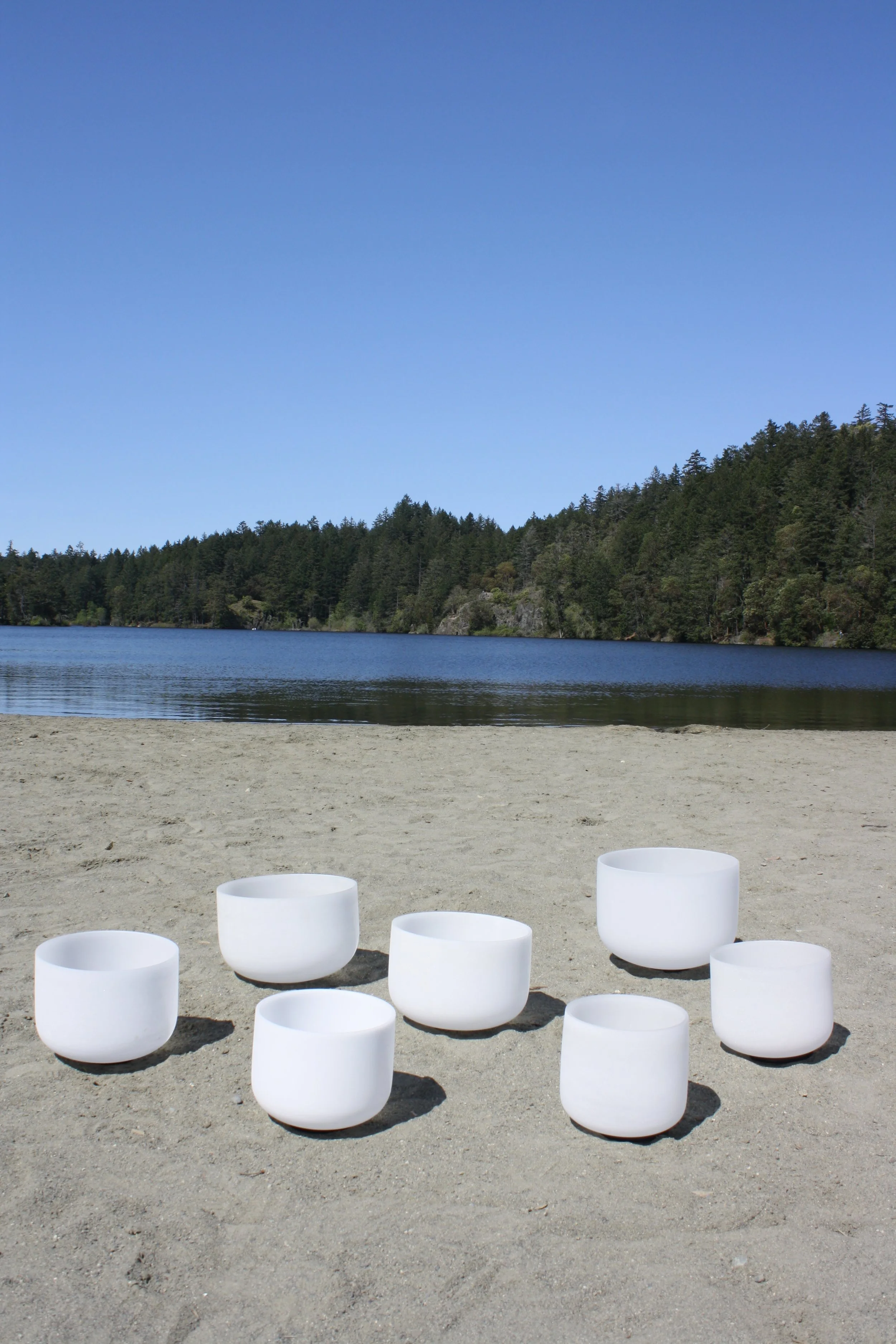 Seven white singing bowls arranged on sandy beach near lake with forested hills and blue sky in the background.