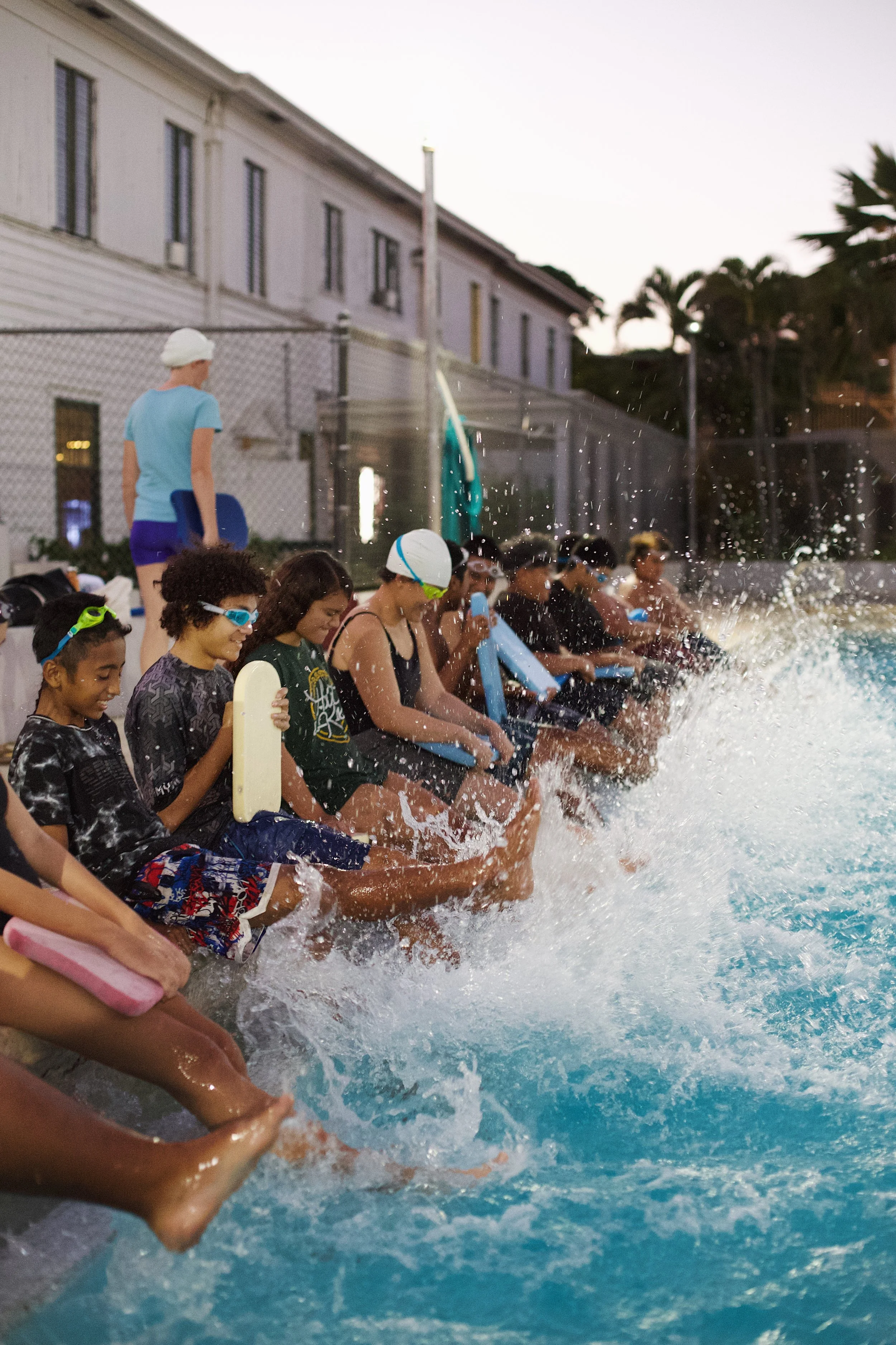 A group of children sitting by the pool's edge, splashing water as they kick their feet in the water. Some are wearing swim goggles and holding kickboards, with an attendant standing nearby.