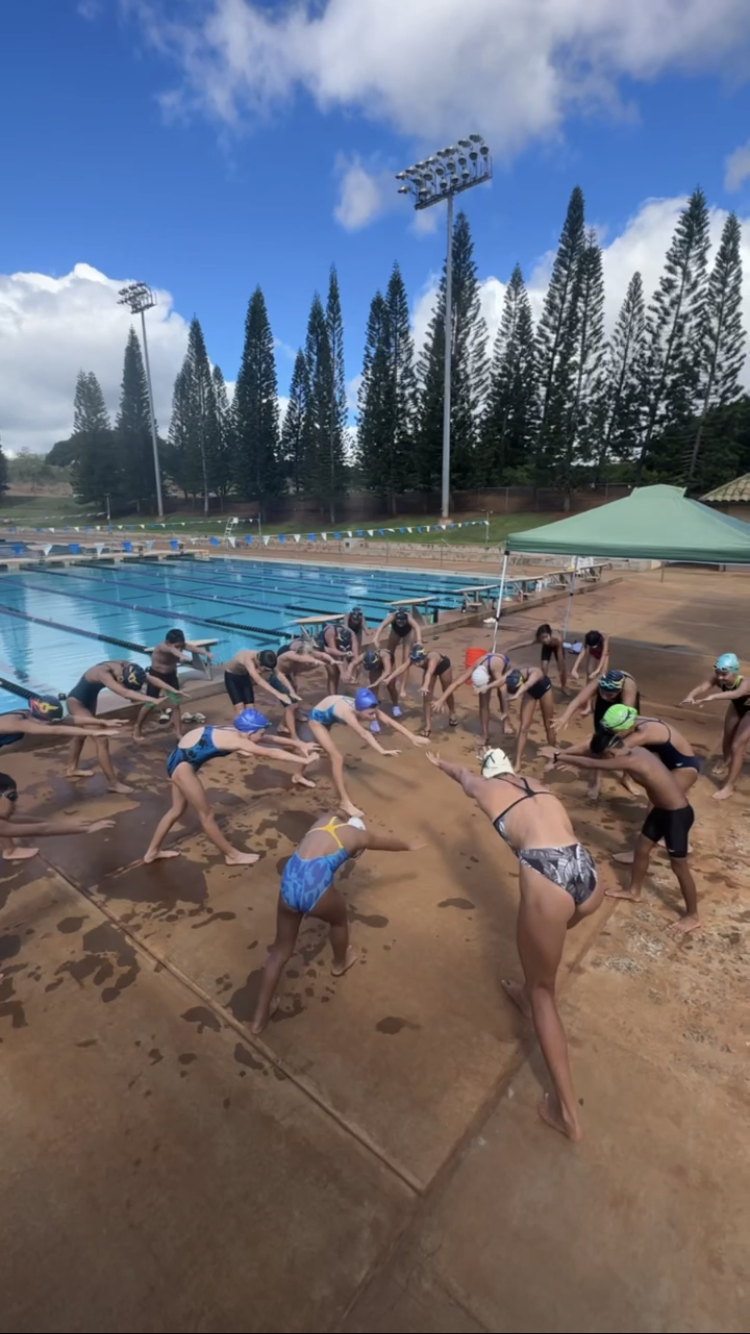 Swimmers doing warm-up exercises on pool deck at a competitive swimming practice or event, with a swimming pool, tall trees, and floodlights in the background.