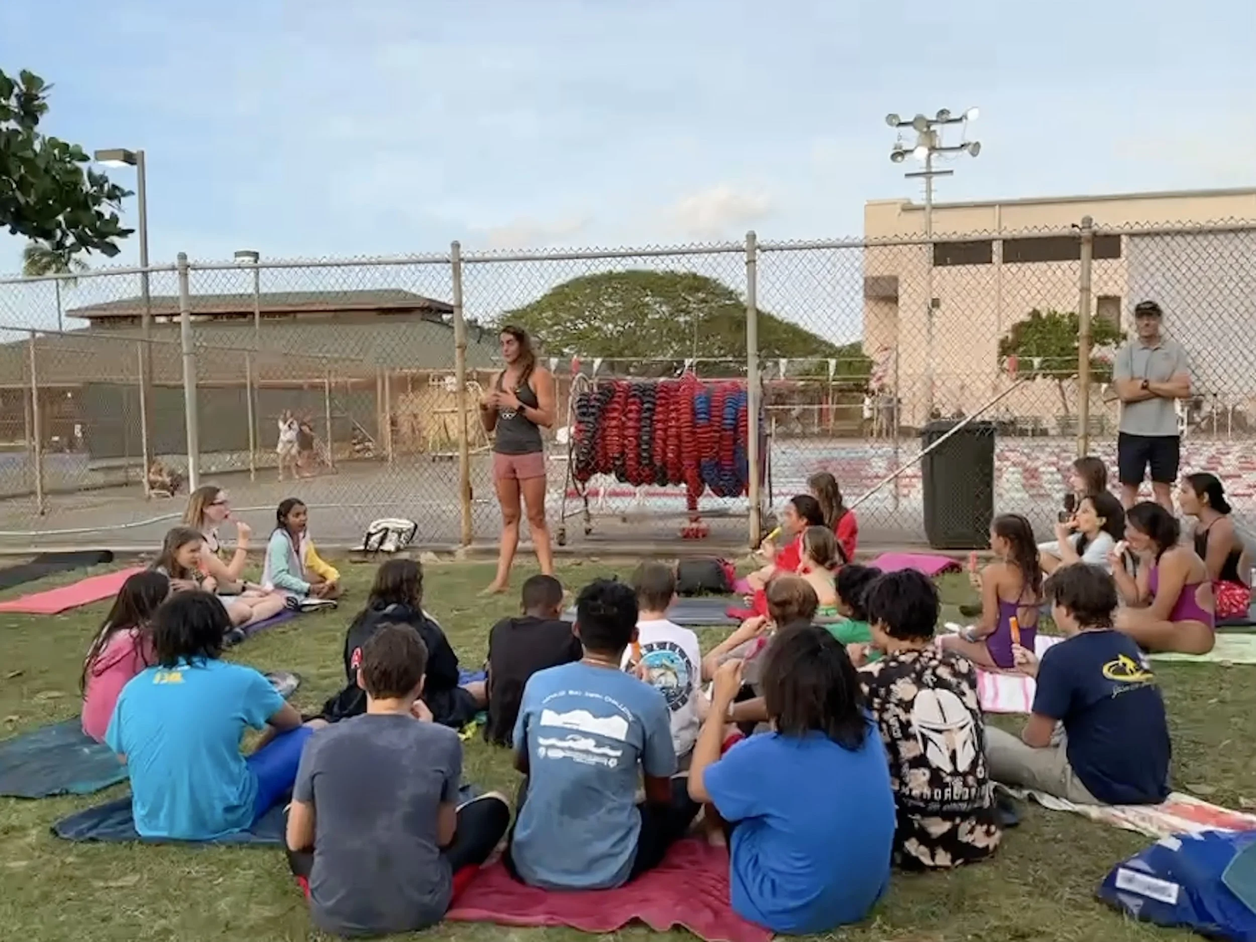 A group of children and teenagers sit on mats on the grass, listening to a woman standing and speaking in an outdoor setting near a fenced area with a tennis court. There are backpacks and other items around them, with some people seen in the backgro