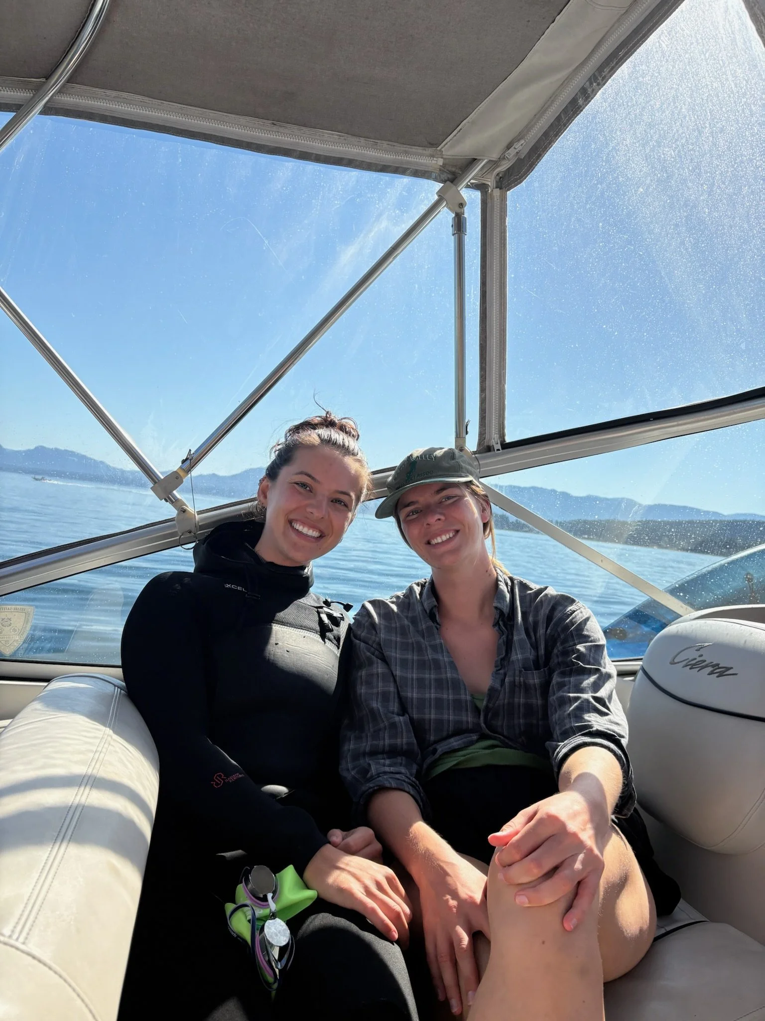 Two women sitting close together on a boat with ocean and mountains in the background, smiling at the camera.