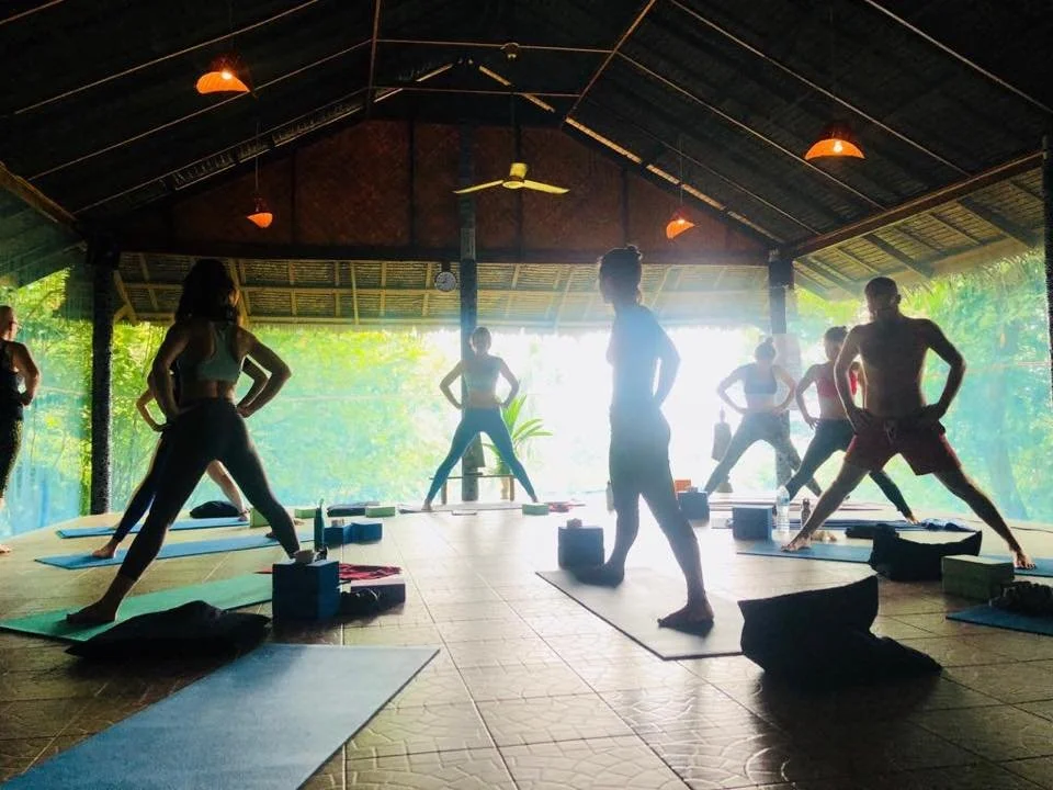 A group of people practicing yoga on mats in an open-air, thatched-roof pavilion with sunlight streaming in from behind.