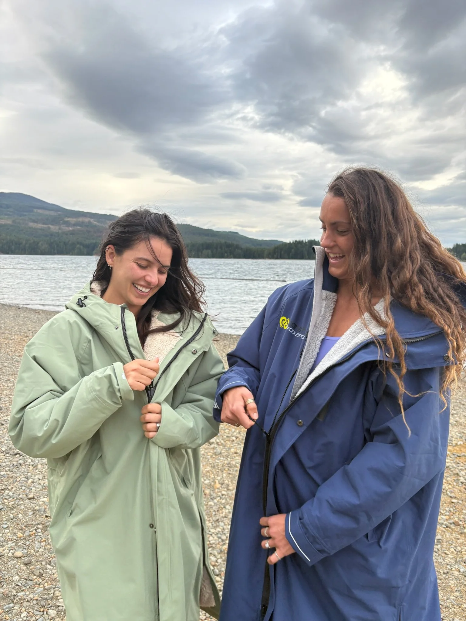 Two women smiling and laughing on a rocky rivershore in weatherproof jackets, with a body of water and mountains under cloudy skies in the background.