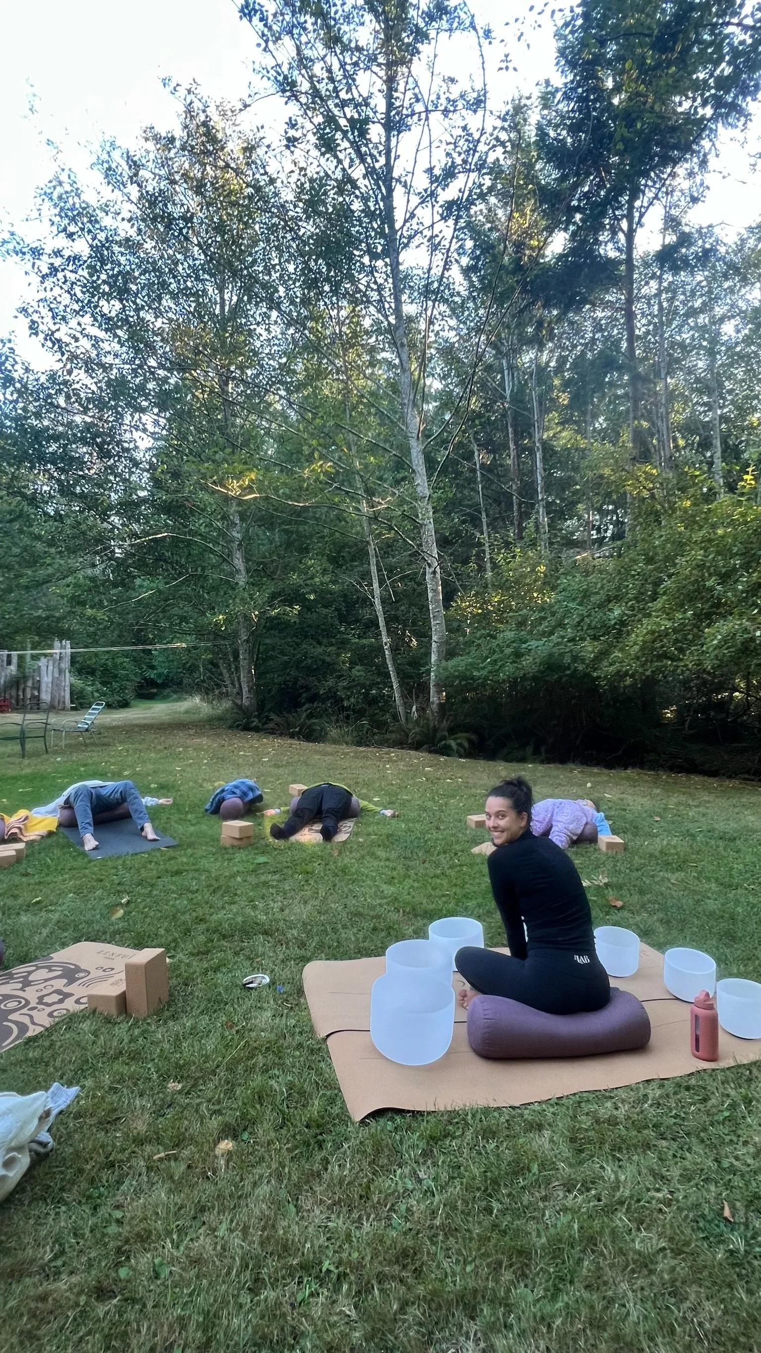 People participating in an outdoor yoga or meditation session on the grass with trees in the background. Several individuals are in a child's pose, and a woman is sitting on a cushion, smiling toward the camera, with crystal singing bowls around her.