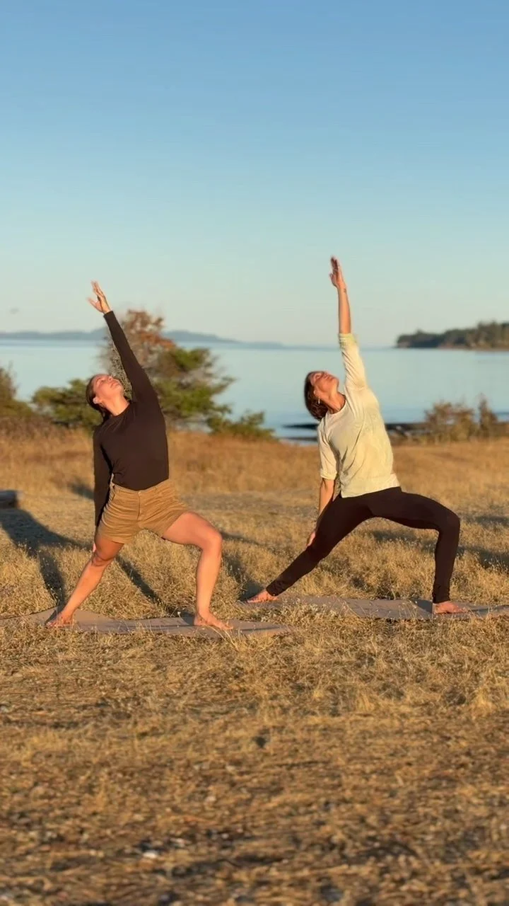 Two women practicing yoga outdoors on mats near a lake, performing a side stretch pose during sunset.