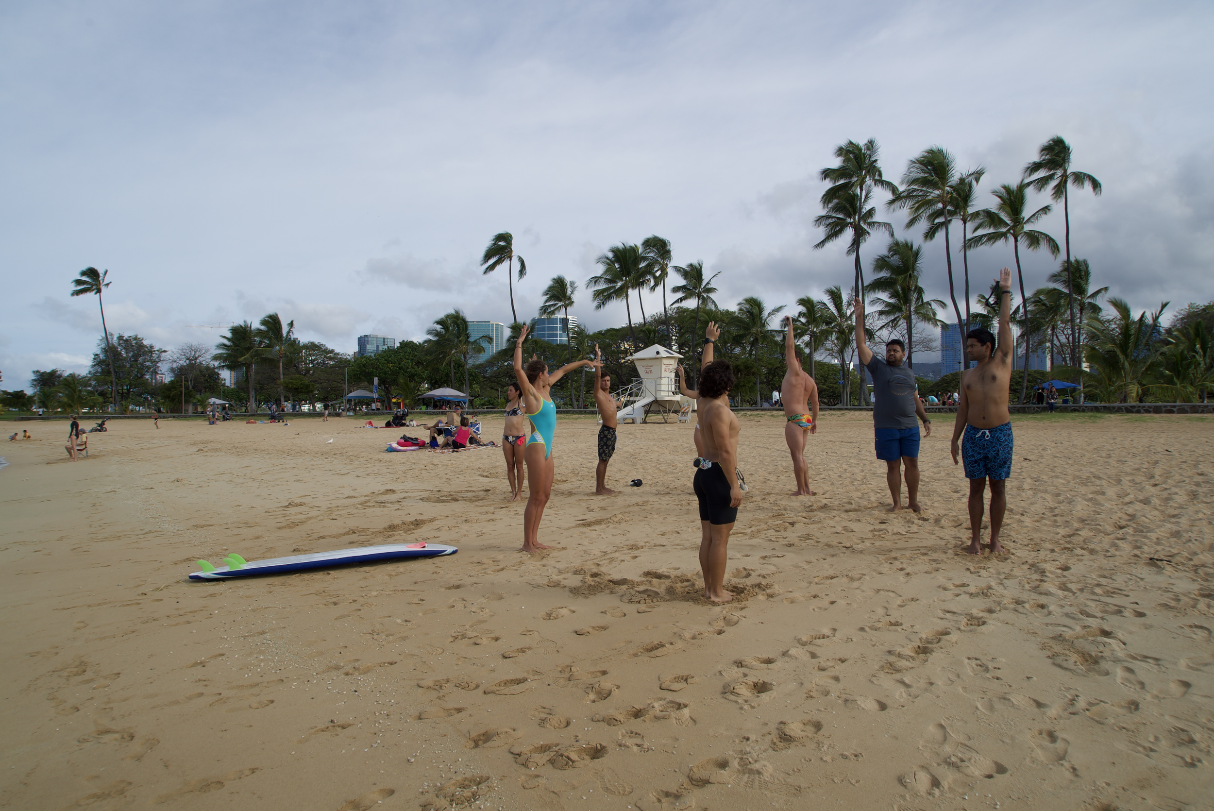 A group of people practicing yoga on a sandy beach with palm trees and city buildings in the background.
