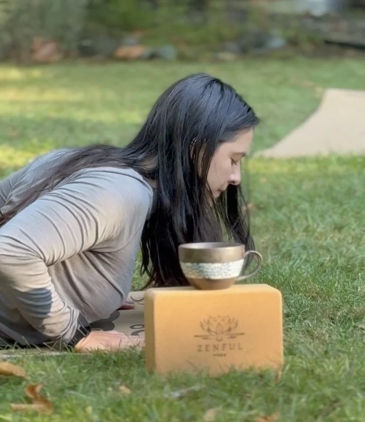 A woman meditating outdoors on grass, with a large cup and a wooden block engraved with 'ZENFUL' in front of her.