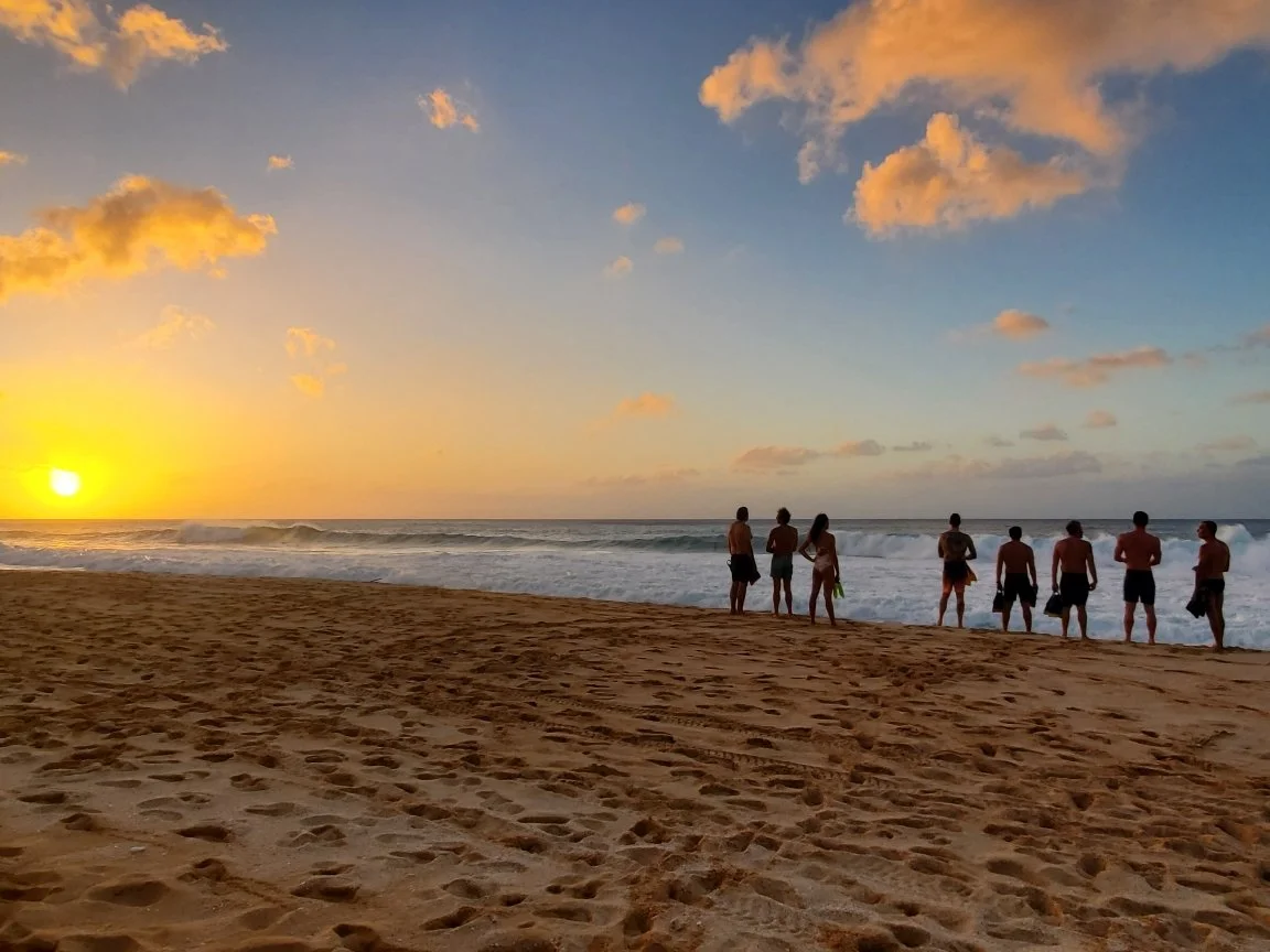 Group of people standing on the sandy beach during sunset, facing the ocean with waves crashing, sky partly cloudy.