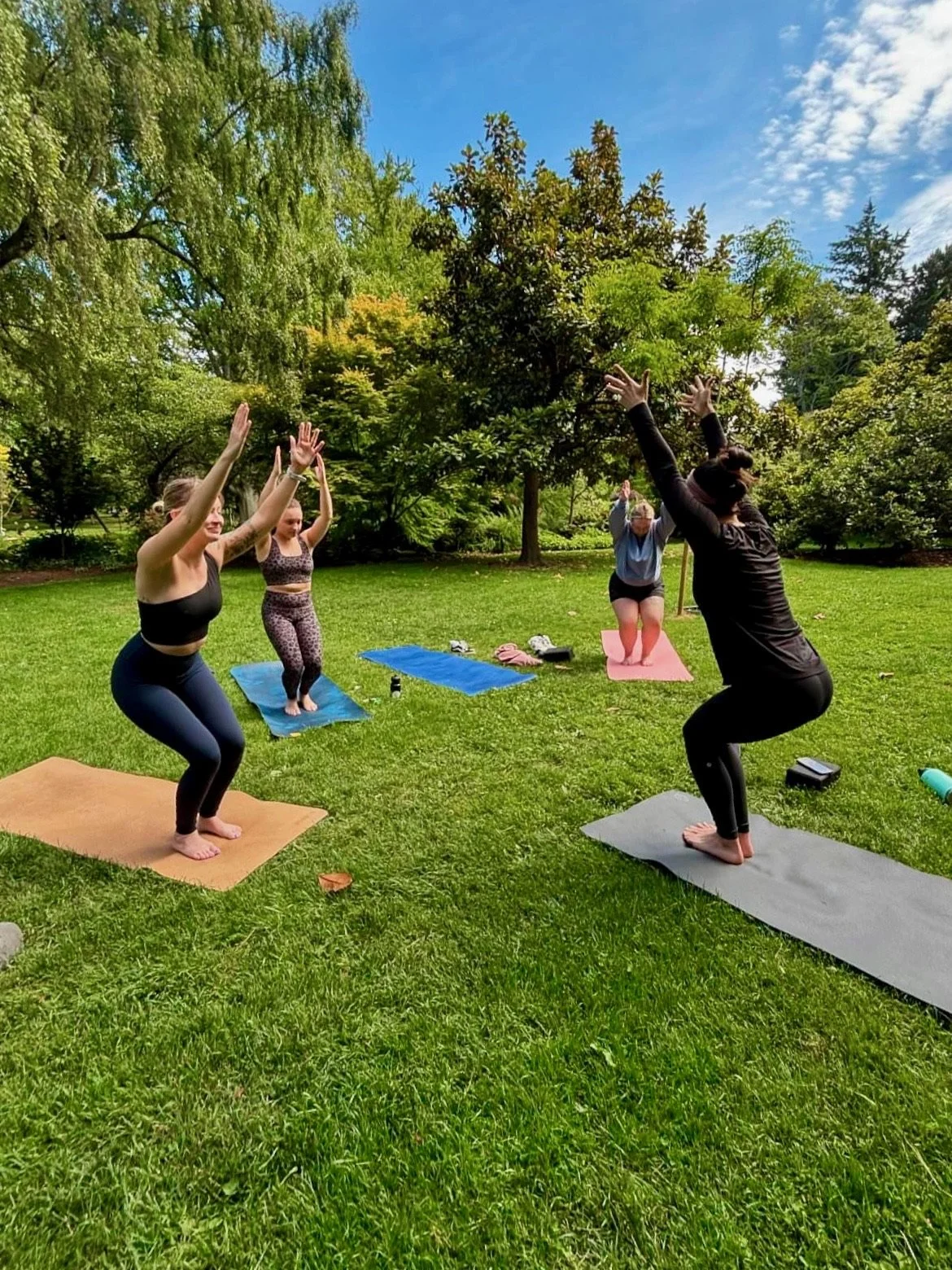 Group of women practicing yoga outdoors on a grassy field surrounded by trees, with a blue sky overhead.