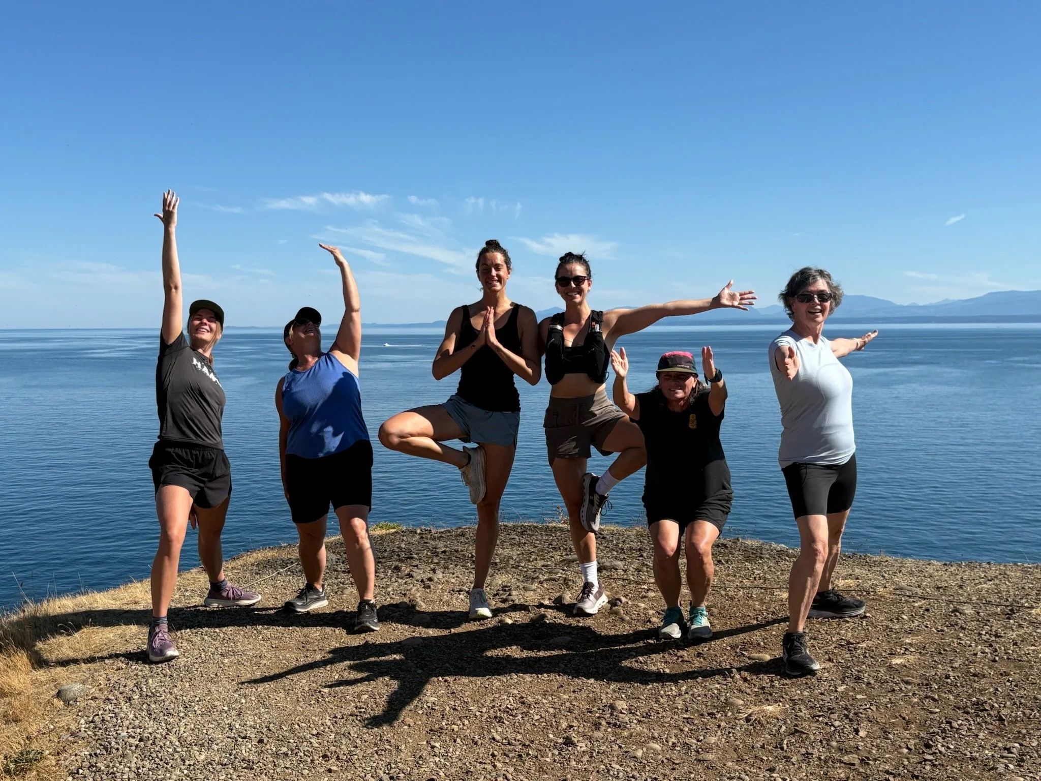 Seven women posing happily on a scenic coastal cliff, with the ocean and mountains in the background under a clear blue sky.