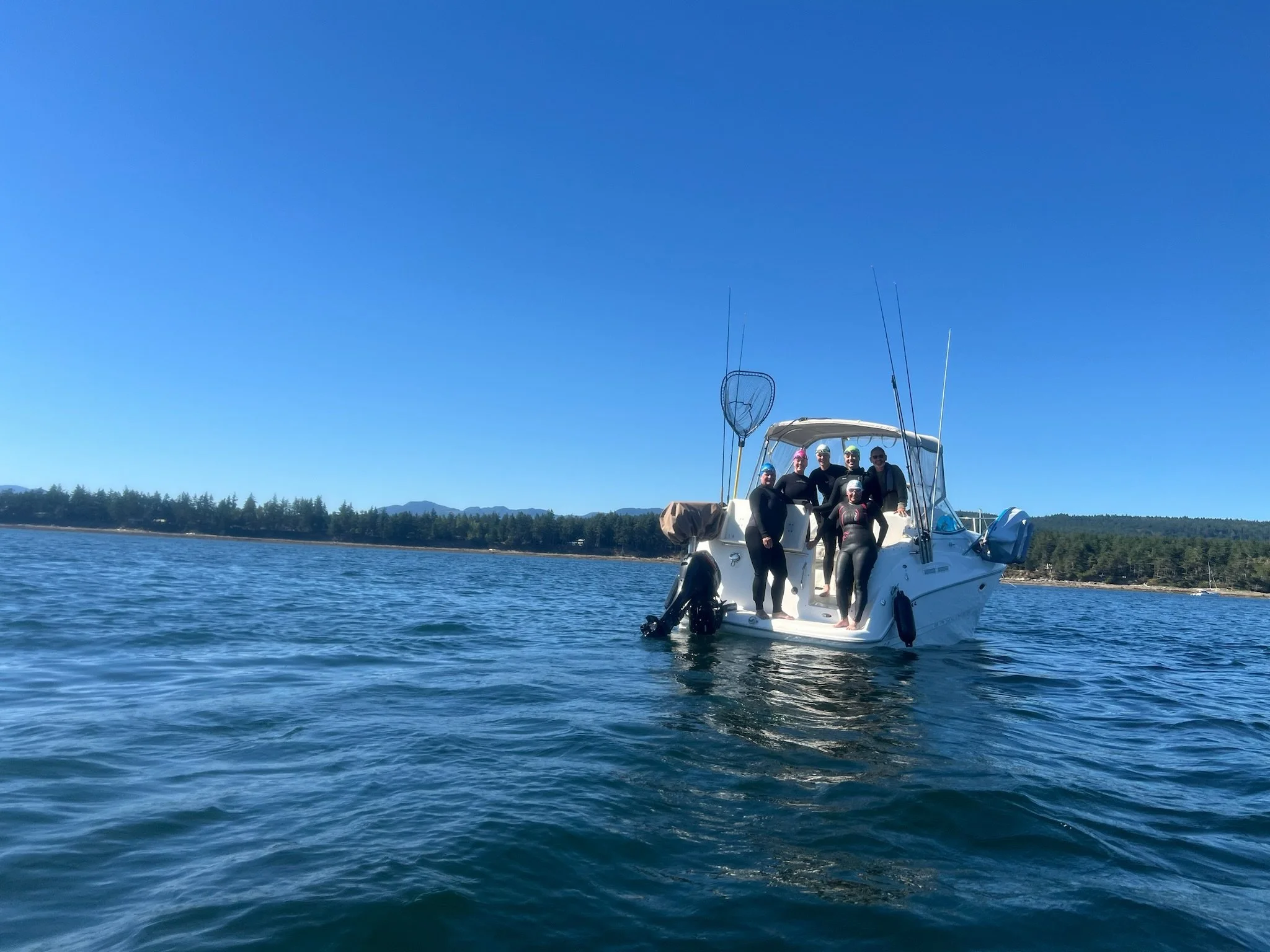 A group of six people in wetsuits on a white boat near the coast with trees and mountains in the background under a clear blue sky.