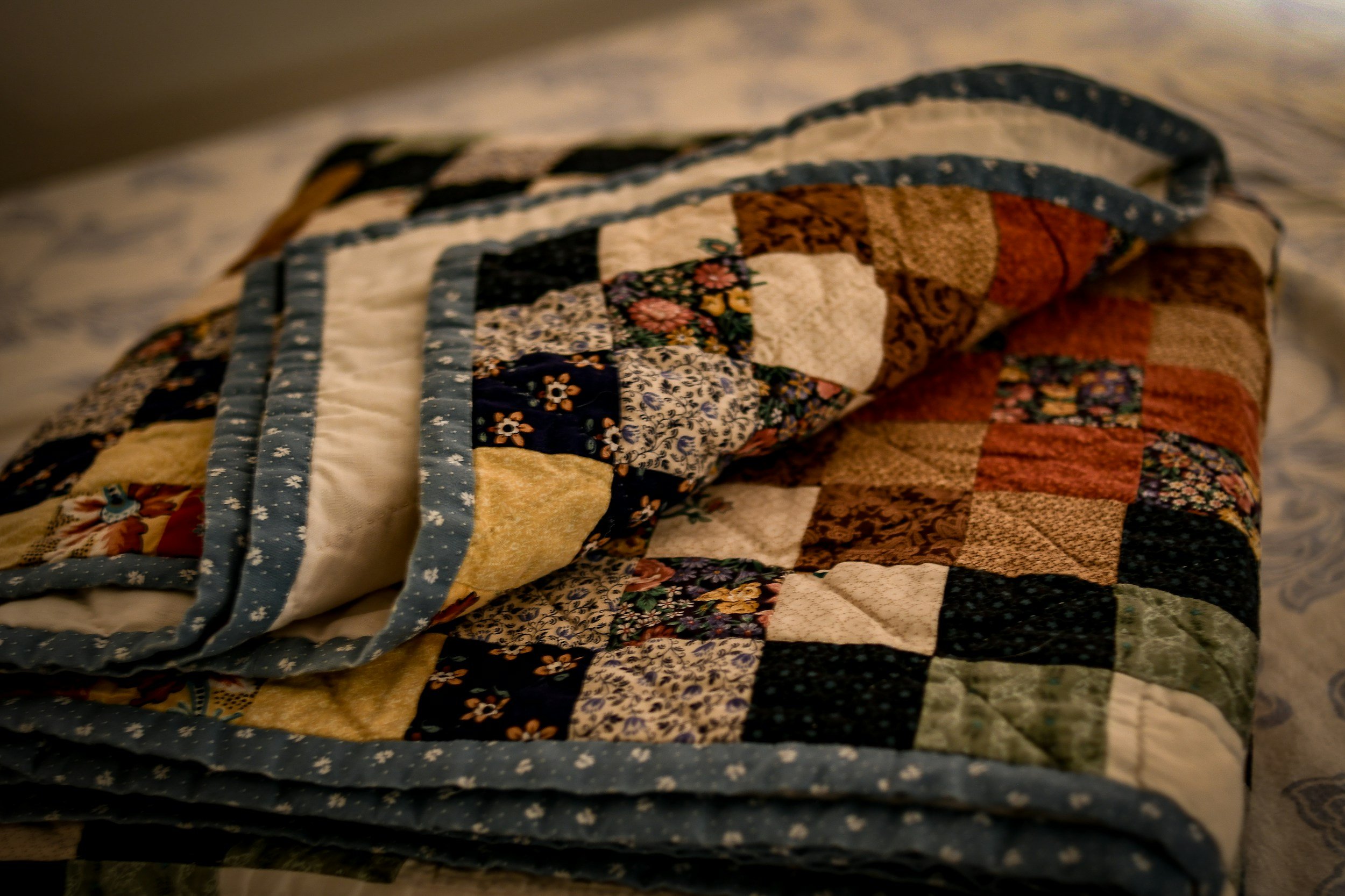 A folded quilt with a patchwork design of various floral and solid fabric squares, bordered with a blue fabric featuring small white flowers, resting on a bed.