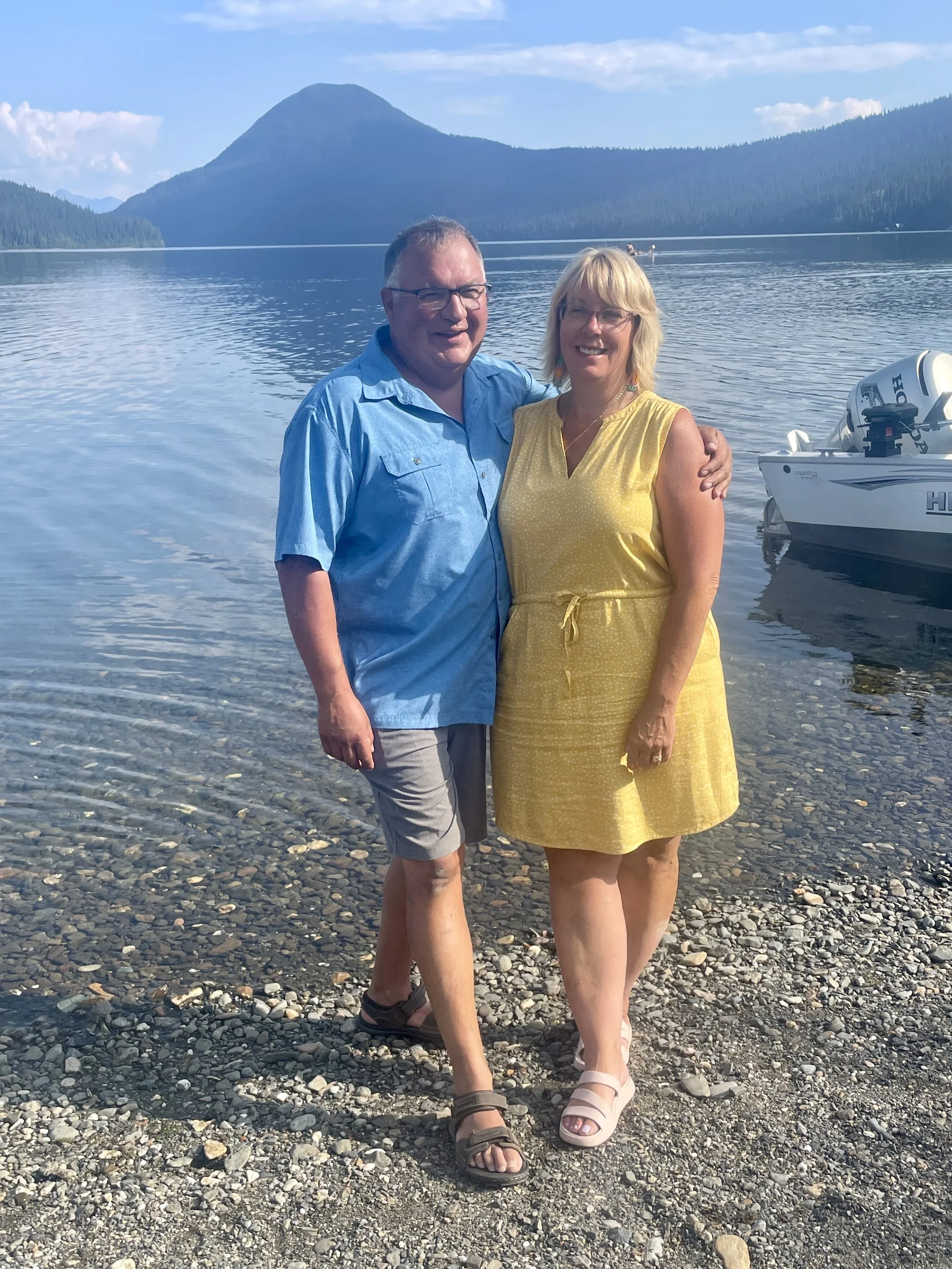 A smiling couple standing on a rocky lakeshore with a mountain and a boat in the background.