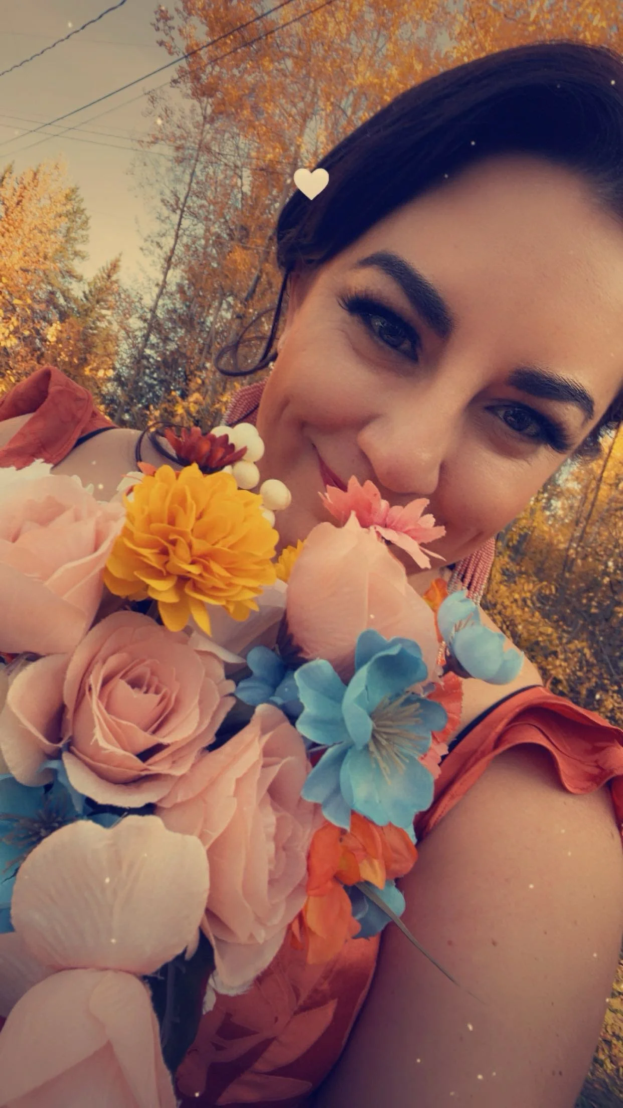 A woman smiling outdoors during autumn, holding a bouquet of pink, yellow, blue, and orange flowers with fall foliage in the background.