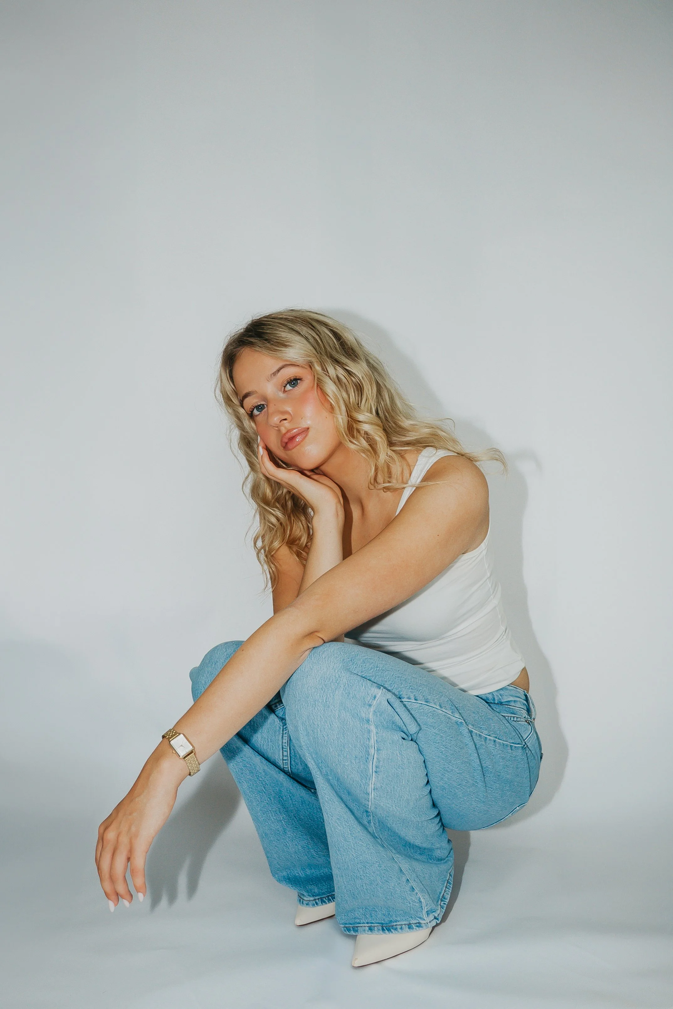 A young woman with blonde wavy hair, sitting on the floor against a plain white background, wearing a white sleeveless top, blue jeans, white shoes, and a gold watch, resting her chin on her hand and looking at the camera.