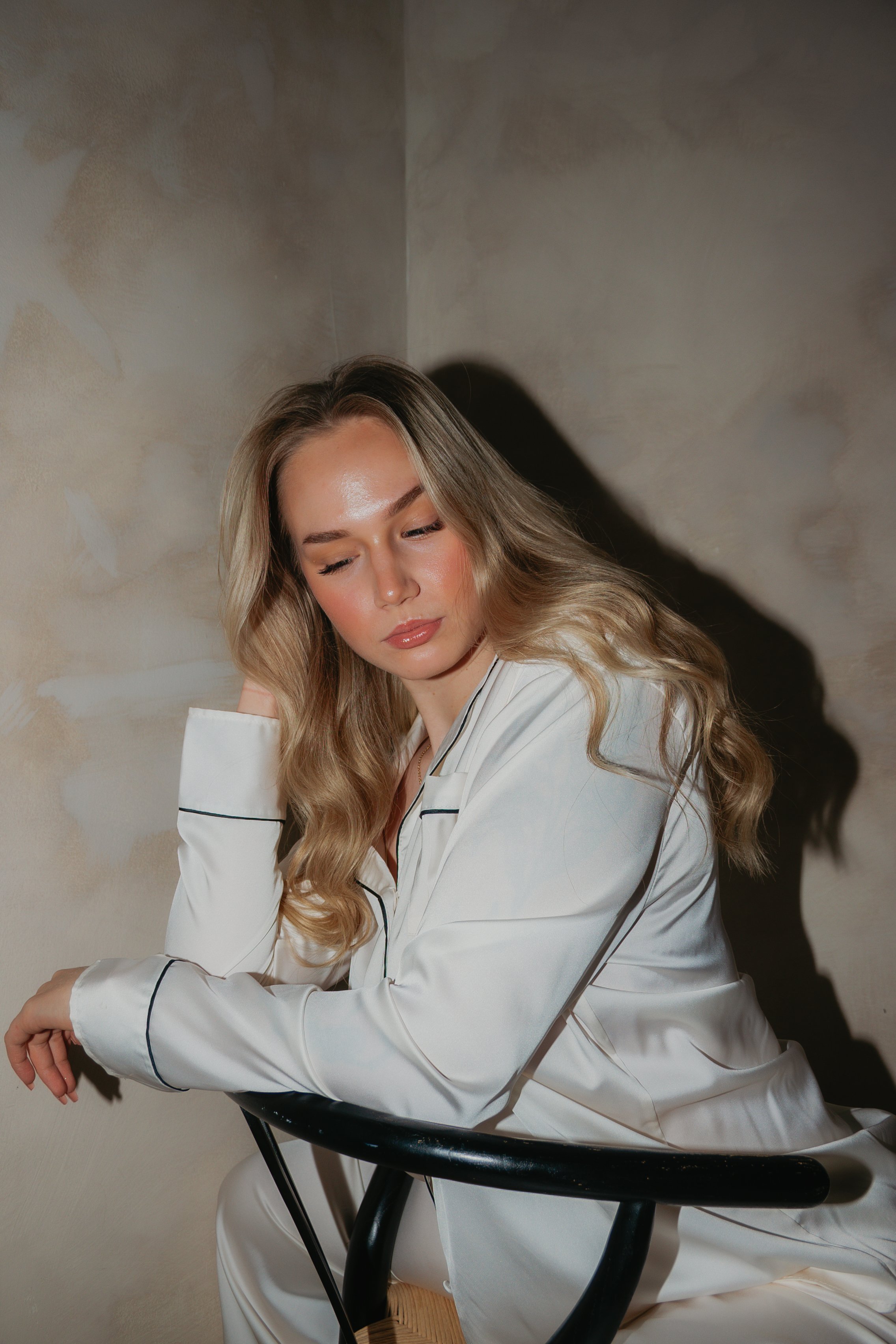 A woman with long blonde hair, closed eyes, and neutral expression, wearing white pajamas, is sitting on a black chair against a beige wall with textured wallpaper.