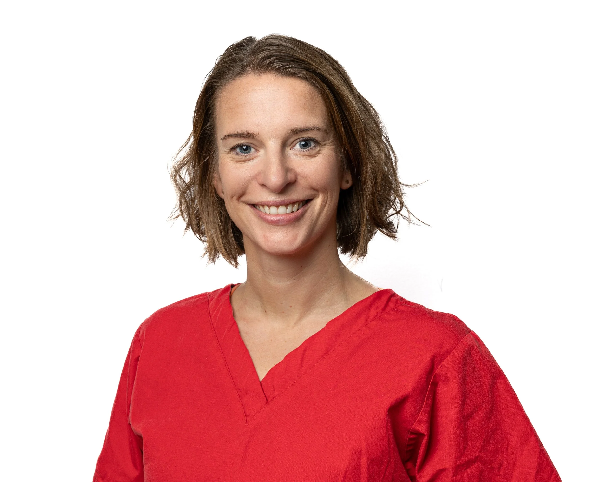 A woman with short wavy brown hair wearing a red medical scrub top, smiling at the camera against a white background.