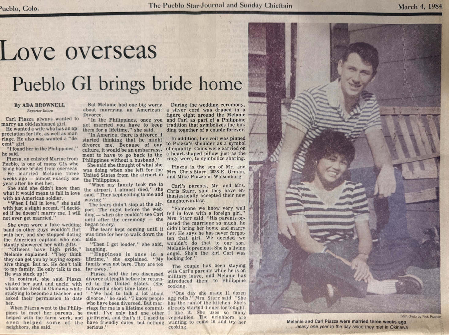 A newspaper clipping from The Pueblo Star-Journal and Sunday Chieftain, dated March 4, 1984. It features a photo of a young man and woman, Melanie and Carl Piazza, both wearing striped shirts. Carl is standing behind Melanie, who is sitting on a wooden stool, with both smiling at the camera. The caption notes they were married three weeks ago in Okinawa, nearly a year after meeting in Okinawa. The article discusses their love story and wedding.