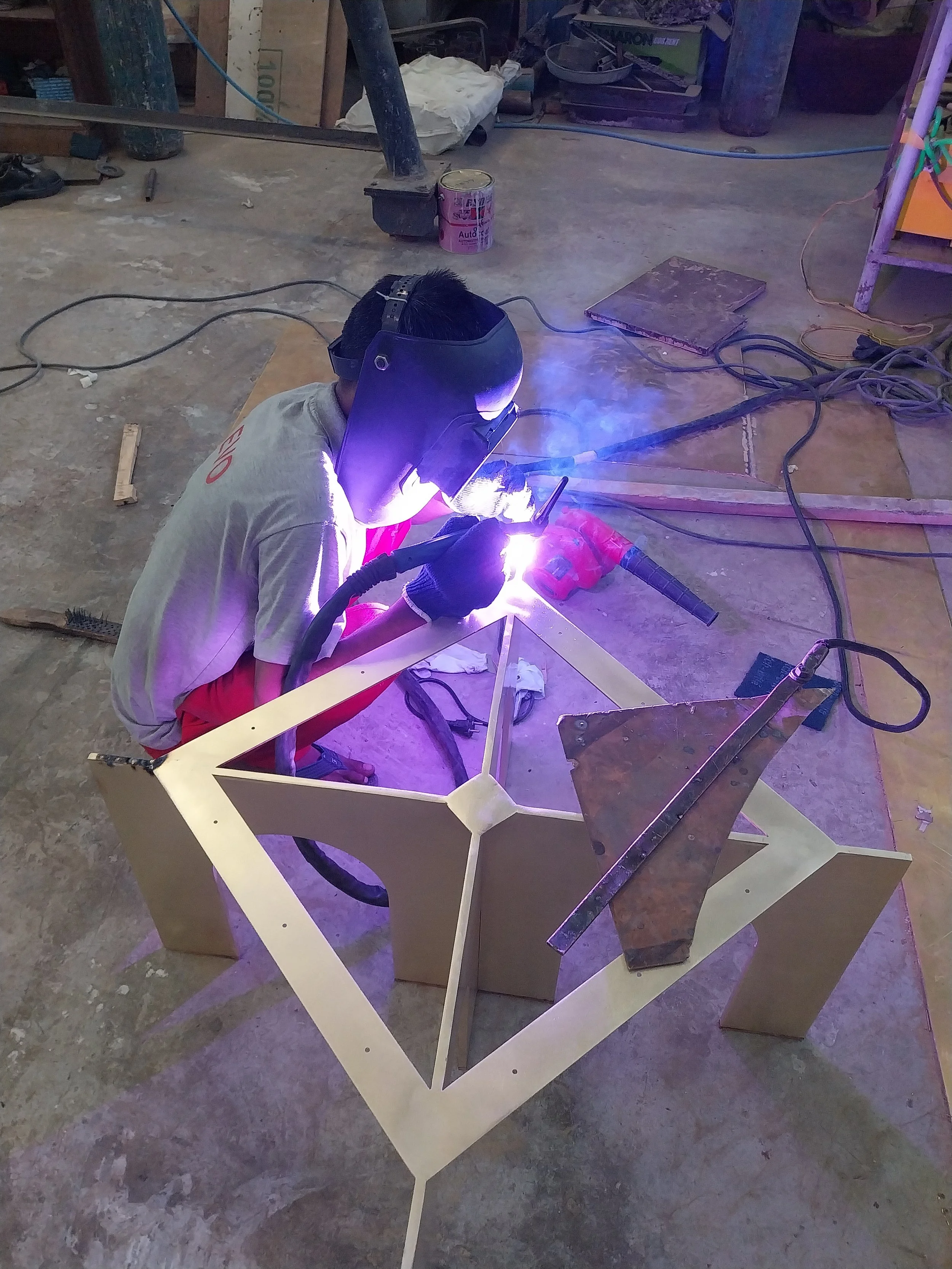 A person is welding a geometric brass table in a workshop, wearing a welding helmet and gloves, with welding equipment and tools around them.