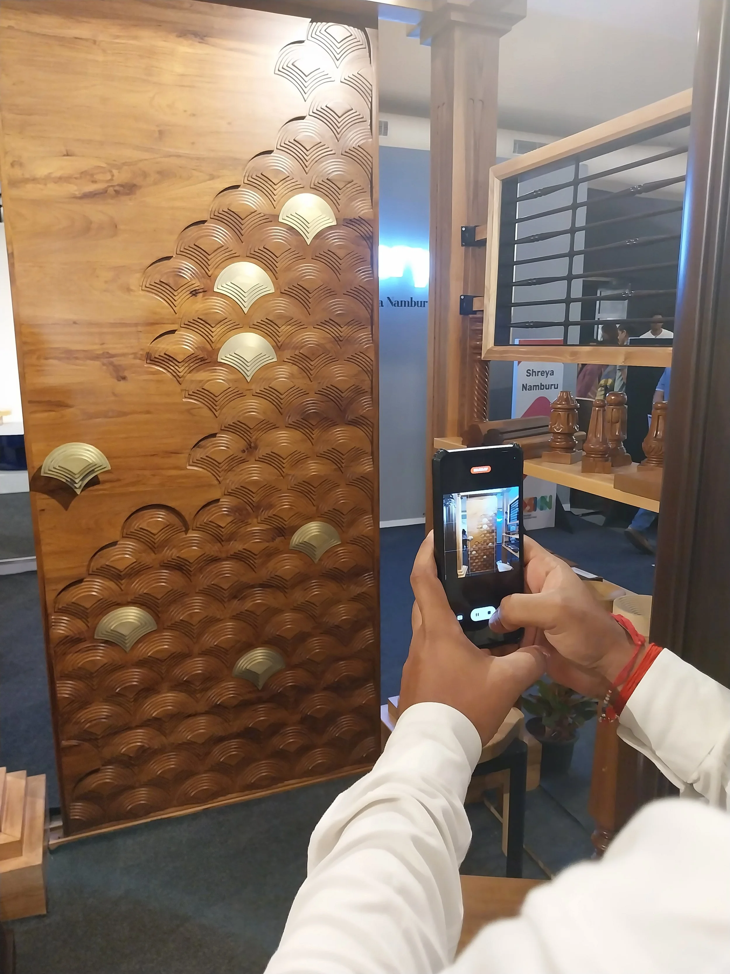 A person taking a photo of a decorative wooden panel with a fan-shaped pattern and brass motif at an indoor event or showroom.