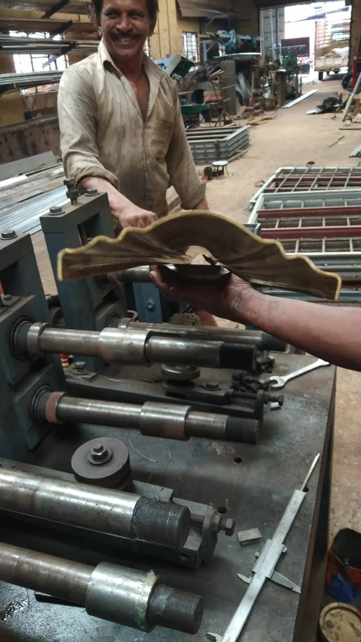 A man smiling in a workshop, holding a brass decorative piece with a wavy edge, while another person hands it over. The workshop has metal parts and tools on the workbench.