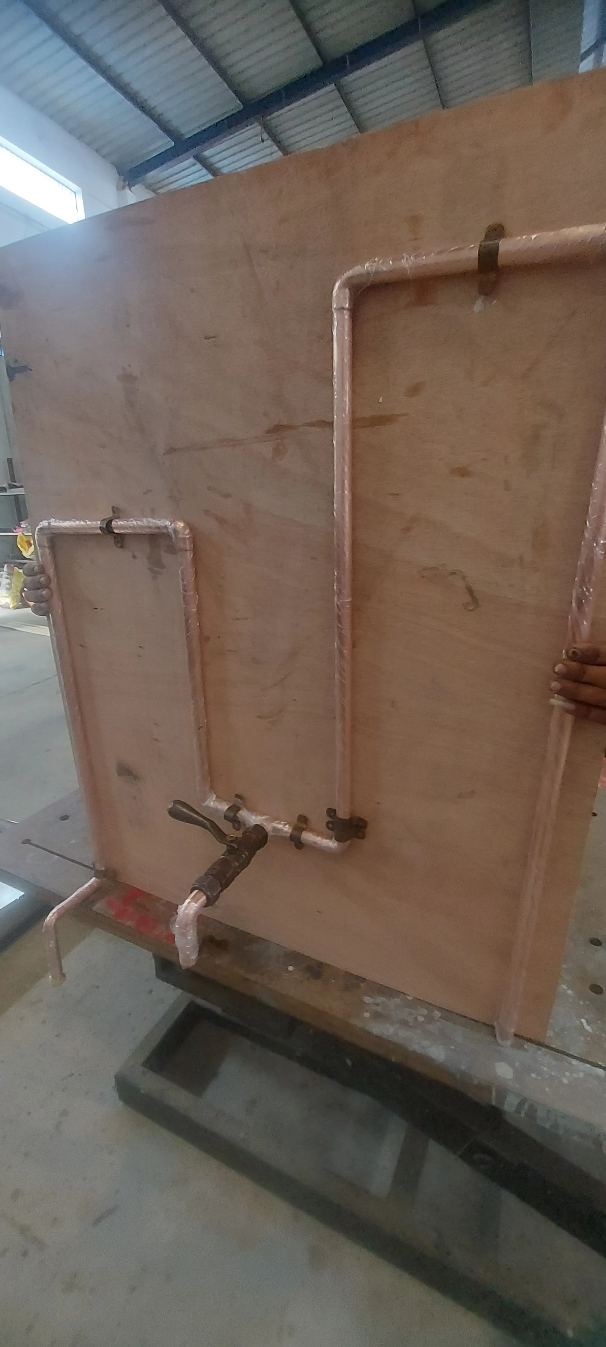 Plywood board with copper piping and brass valve arranged in a pattern and connected to a faucet, likely part of a plumbing or water system setup.