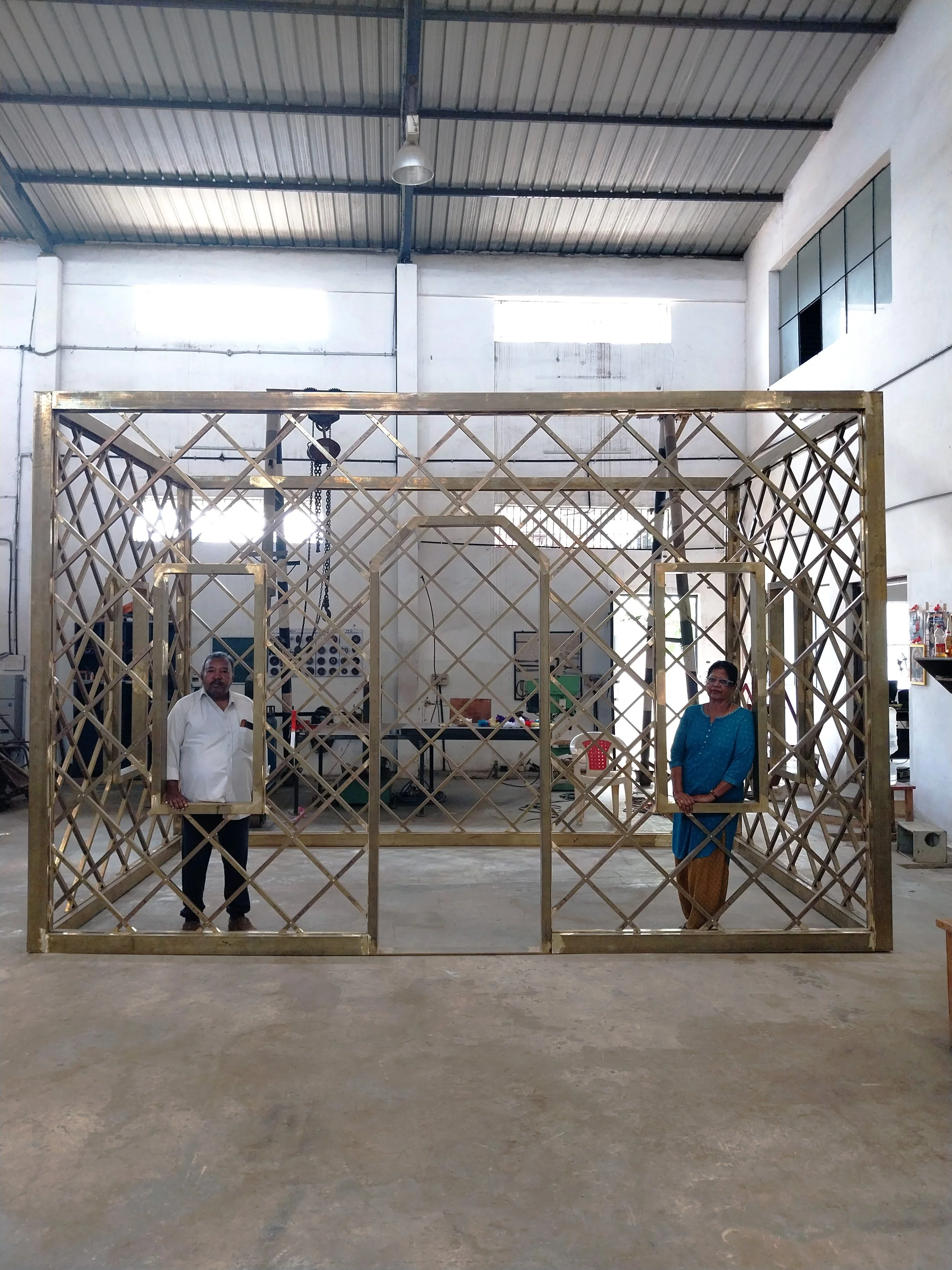 Two individuals, a man and a woman, are standing inside a large, brass lattice structure inside a spacious warehouse or workshop with high ceilings and metal roofing.