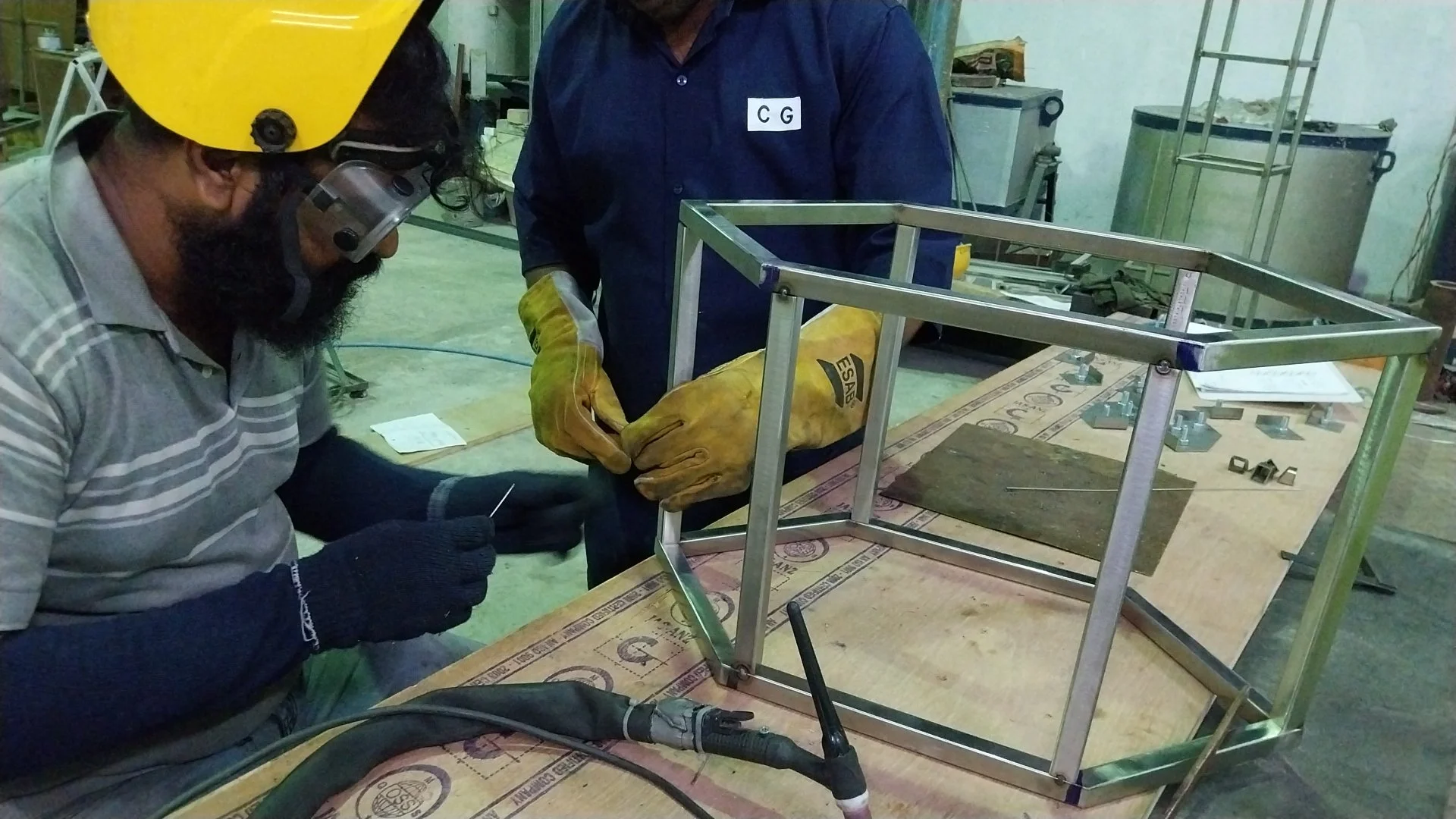 Two men working on a metal frame structure in a workshop, one wearing a yellow helmet and welding gloves, the other with safety glasses and gloves, welding or assembling the frame.