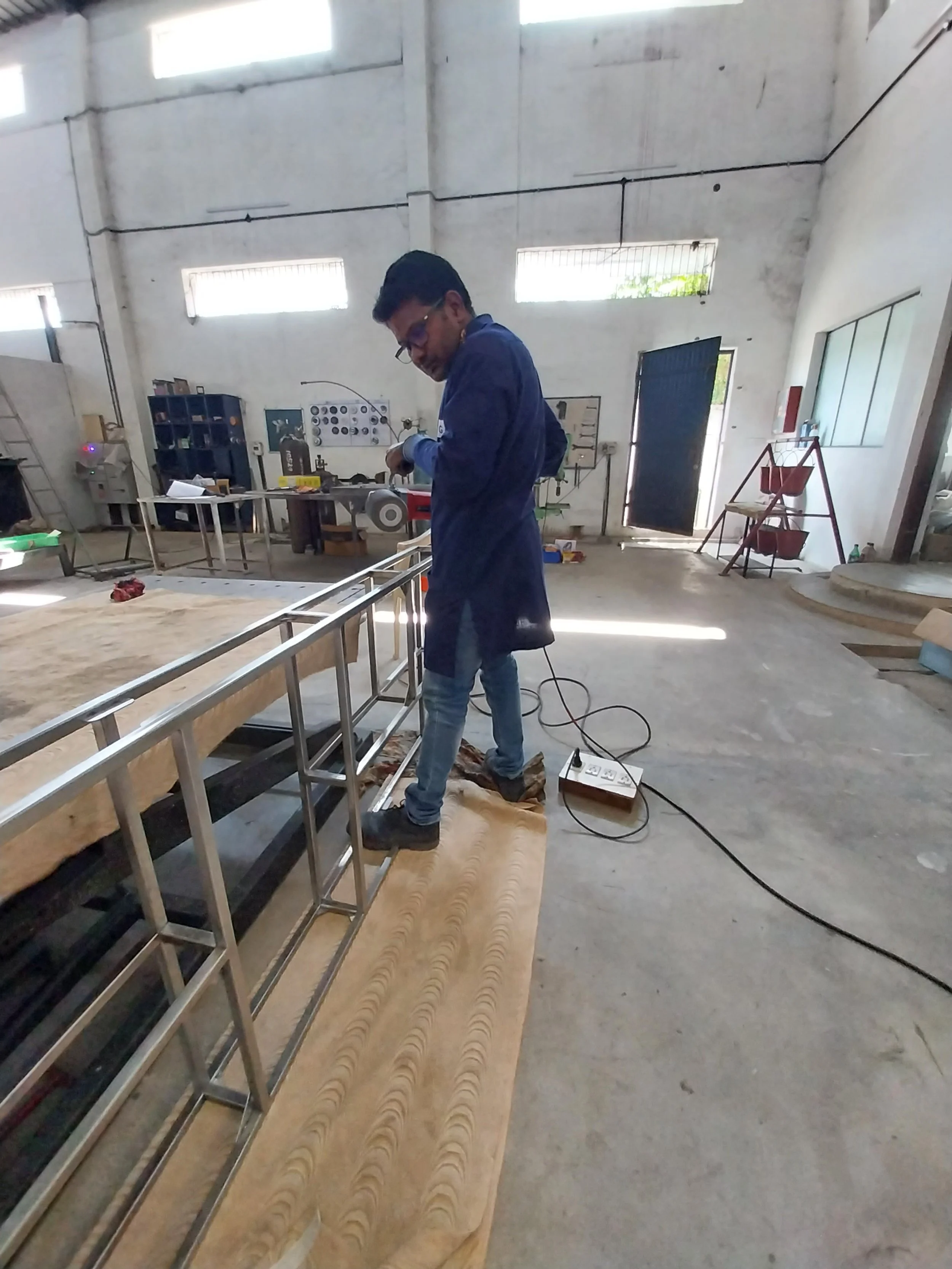 A man working on a metal railing inside a spacious, industrial-style workshop or factory with high ceilings and large windows.