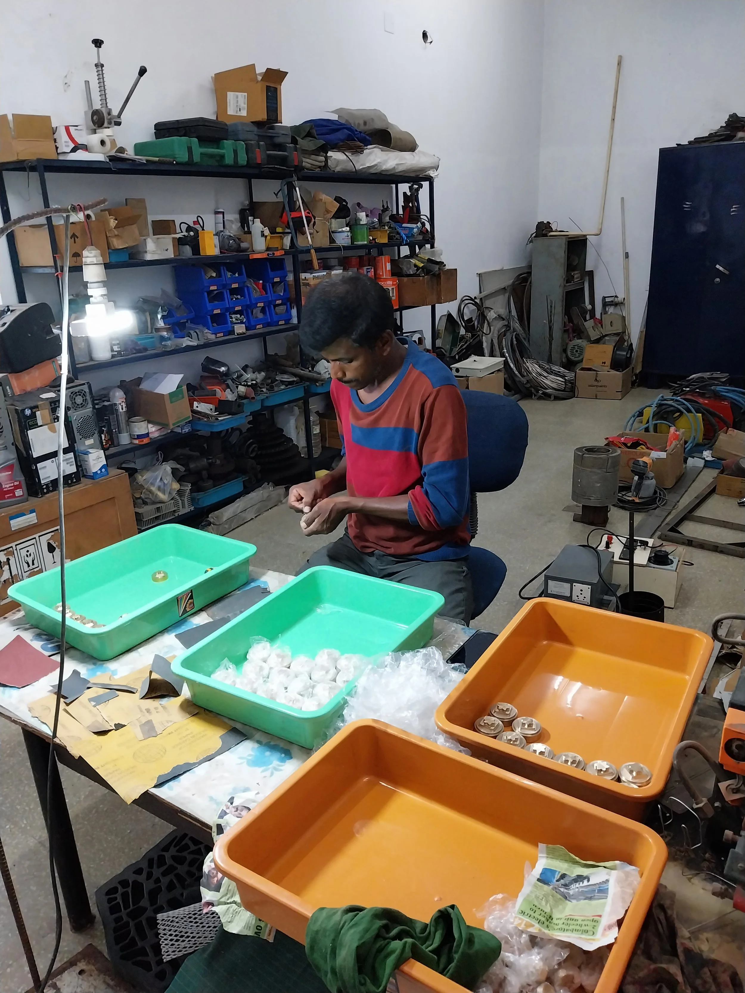 A man is sitting at a cluttered workbench in a workshop, sorting and working with small items. The workspace has trays, tools, and various electronic parts, with shelves filled with boxes, parts, and supplies behind him.