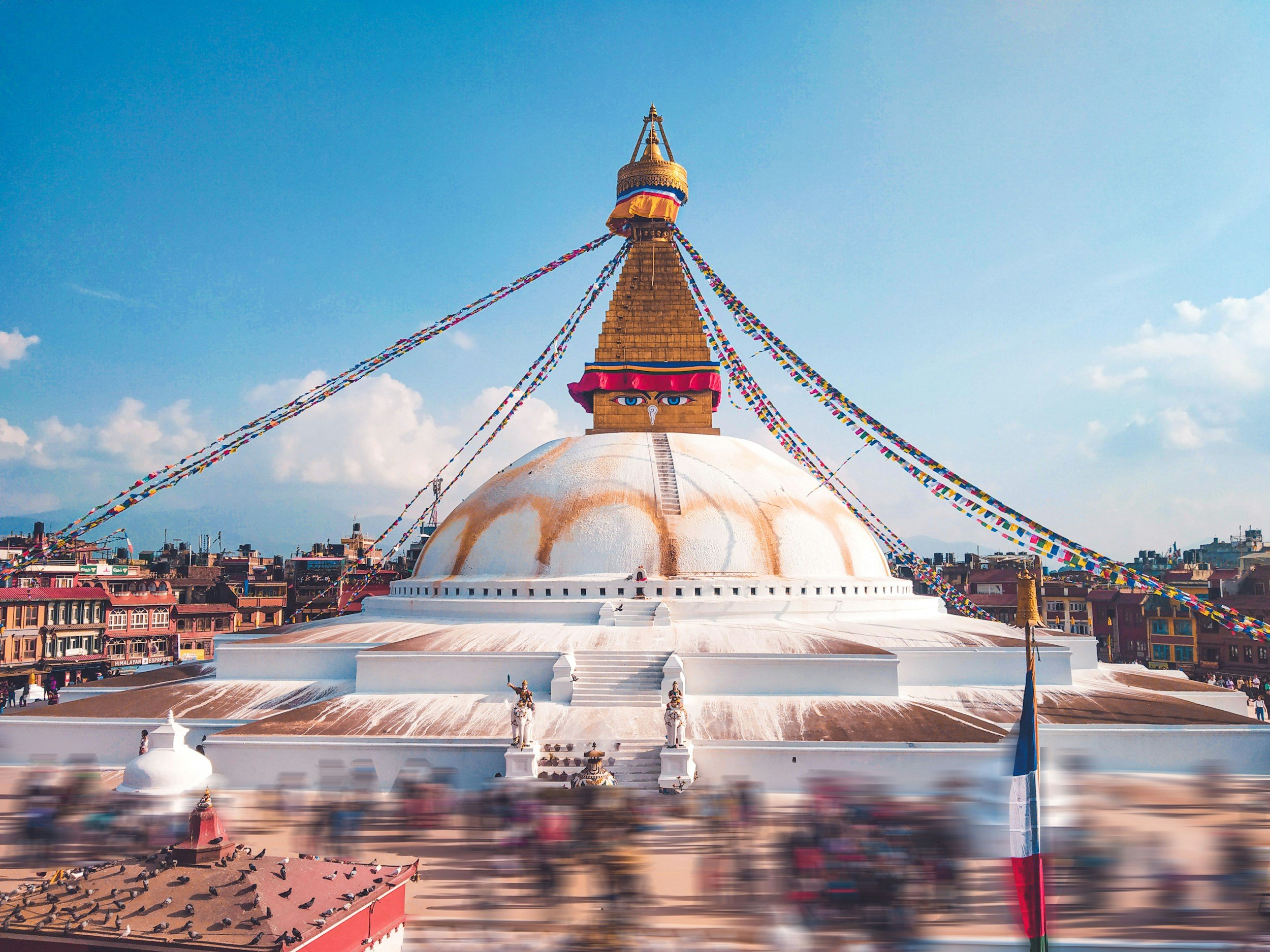 A large Buddhist stupa decorated with prayer flags, with a clear sky and cityscape in the background.