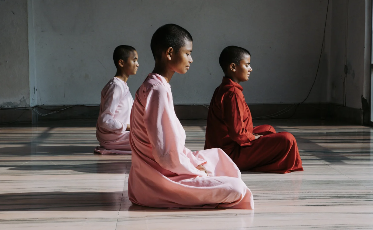 Three young boys or monks sitting cross-legged on the floor in a prayer or meditation pose, dressed in pink and red robes, with a plain wall and window in the background.