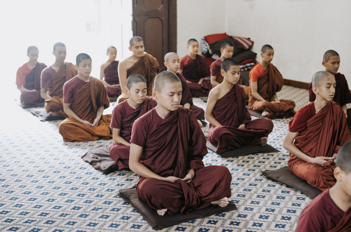 A group of young Buddhist monks meditating in a prayer room, all seated on cushions with closed eyes, practicing mindfulness or prayer.