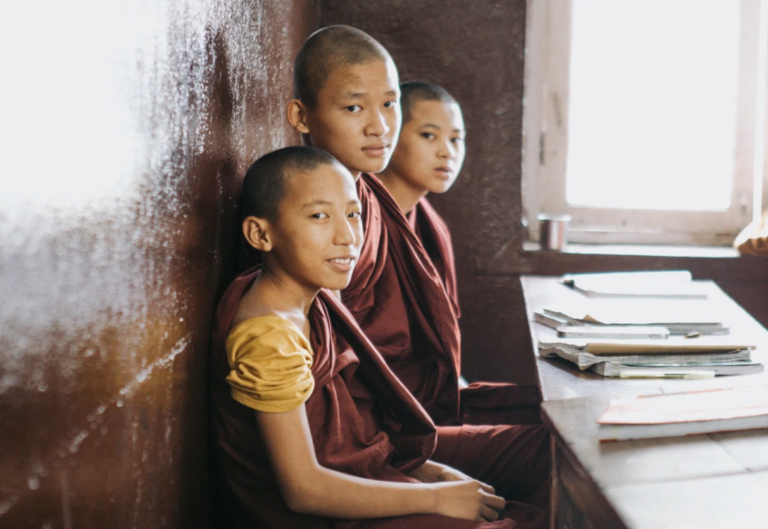 Three young Buddhist monks sitting in a classroom, looking at the camera, with notebooks on the desk.
