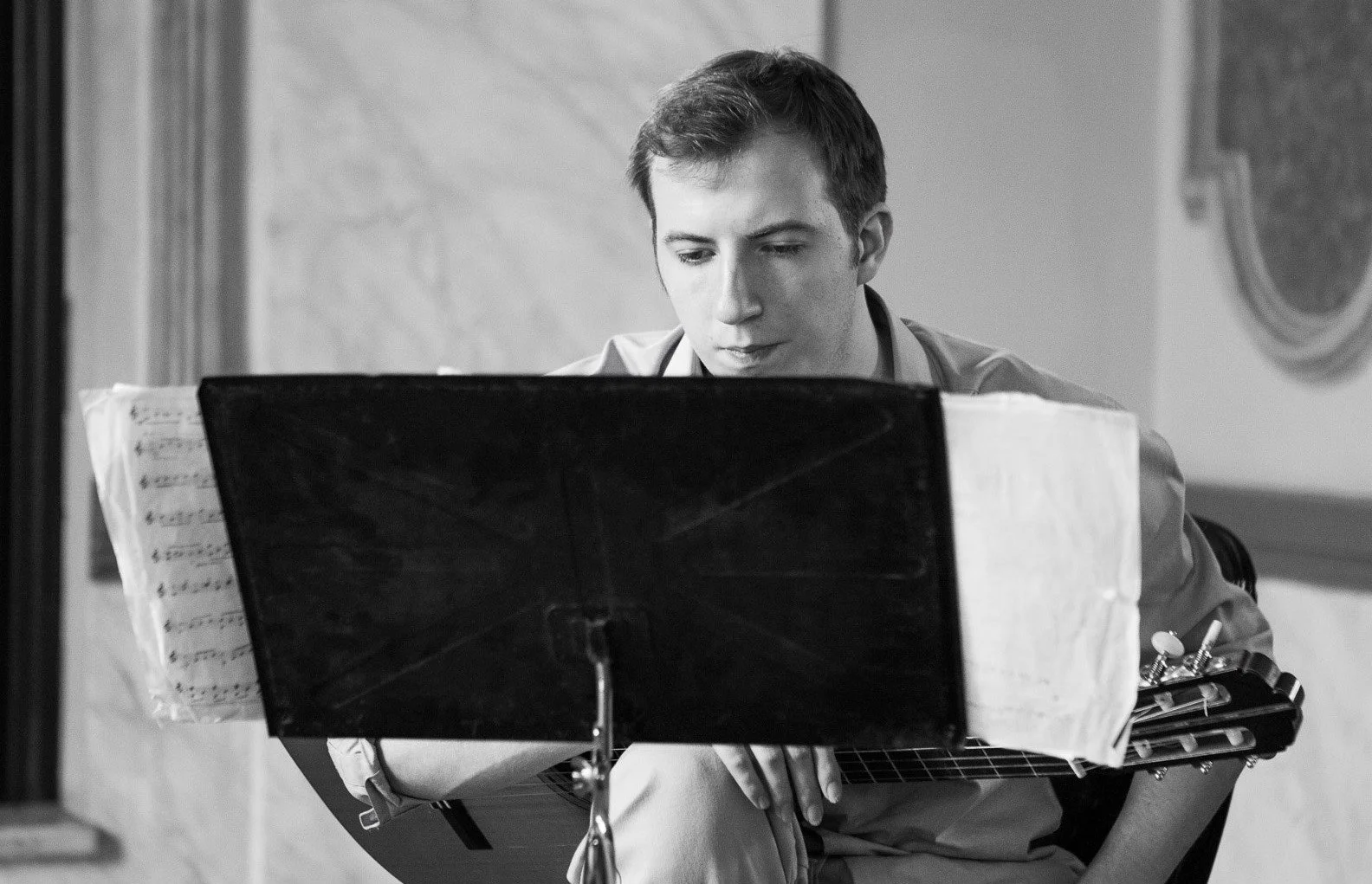 A young man with short hair playing an acoustic guitar, looking down at sheet music on a music stand.