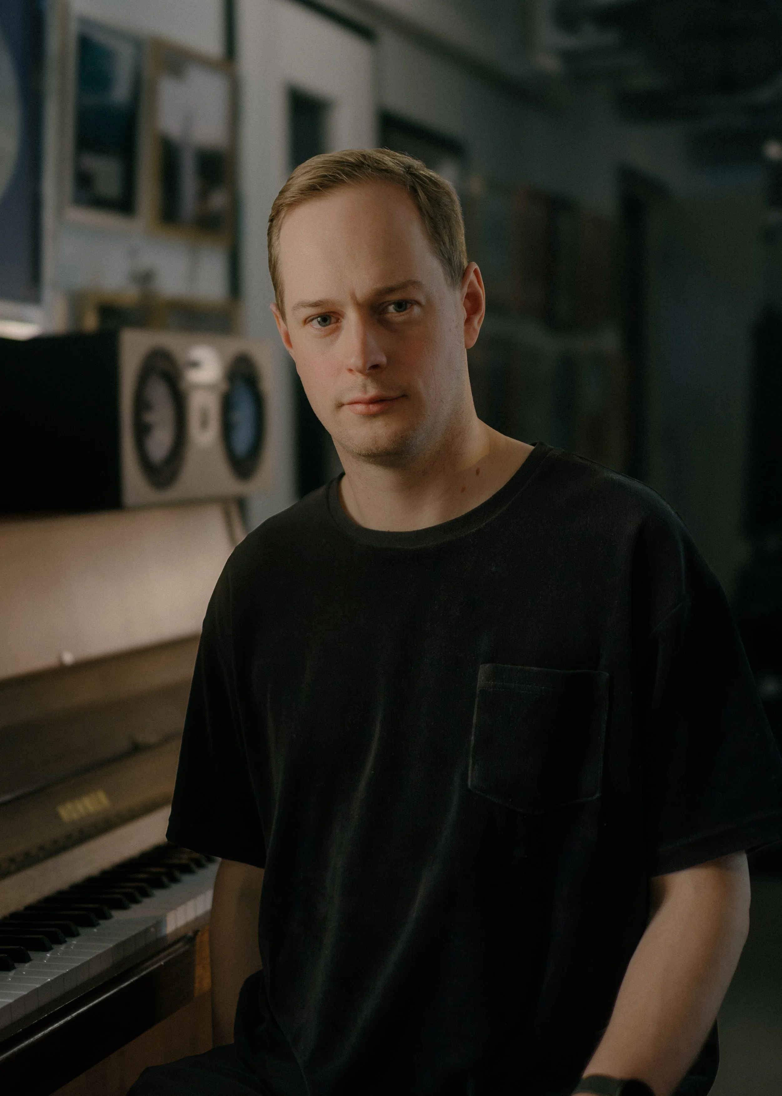A young man with short light brown hair and fair skin stands beside a piano, looking at the camera with a neutral expression. He is wearing a black T-shirt.