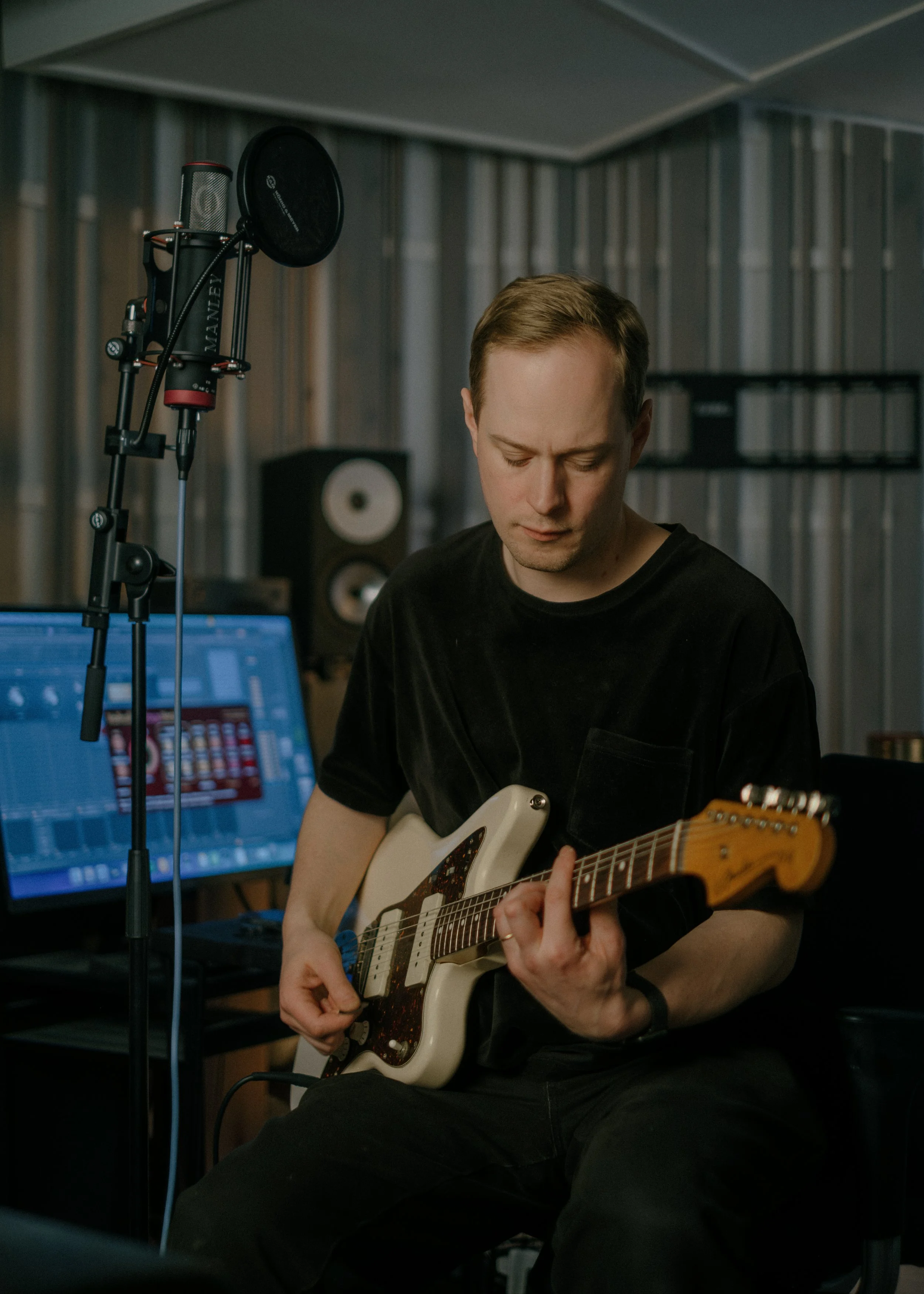A man playing an electric guitar in a recording studio with sound equipment and computer monitors in the background.